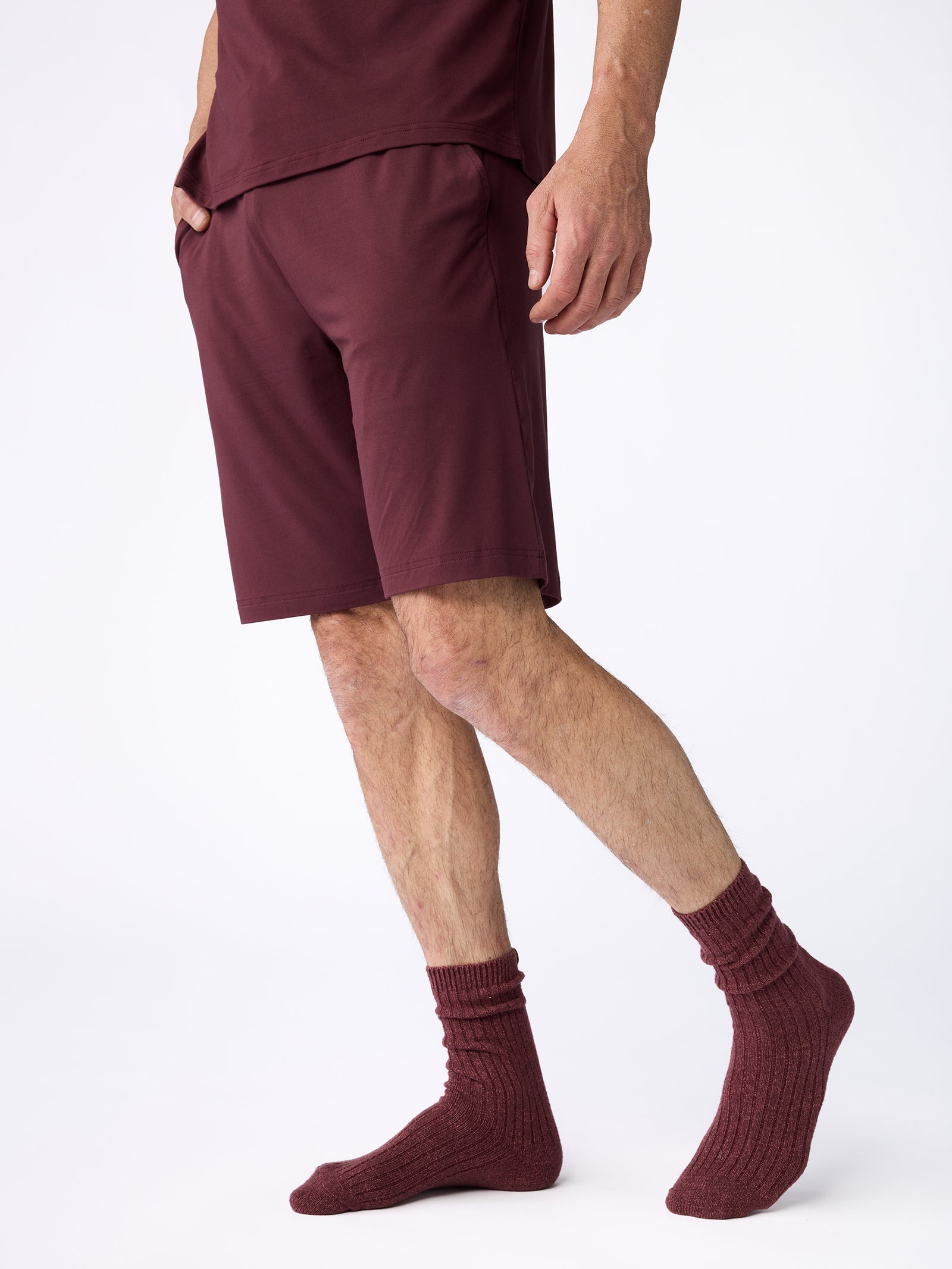 A person is dressed in a coordinated set of burgundy attire, featuring Cozy Earth's Men's Stretch-Knit Bamboo Pajama Short, along with a t-shirt and socks. The scene is set against a simple white backdrop, and the person's face remains hidden from view. 