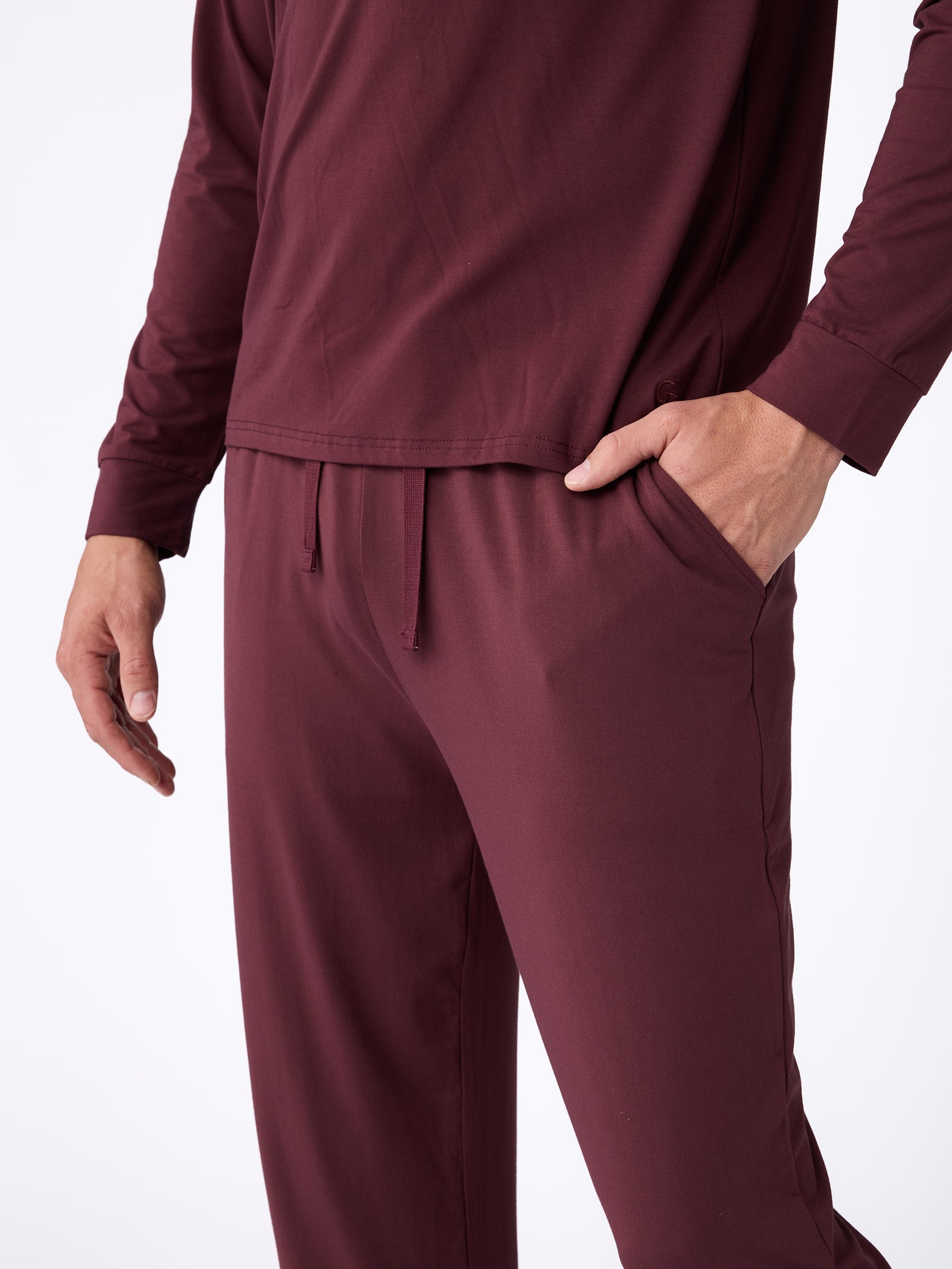 A person wearing a matching maroon long-sleeve shirt and the Men's Stretch-Knit Bamboo Pajama Pant by Cozy Earth. The outfit is casual with a drawstring waistband, and the person's right hand is in the pants pocket. The background is plain white. 