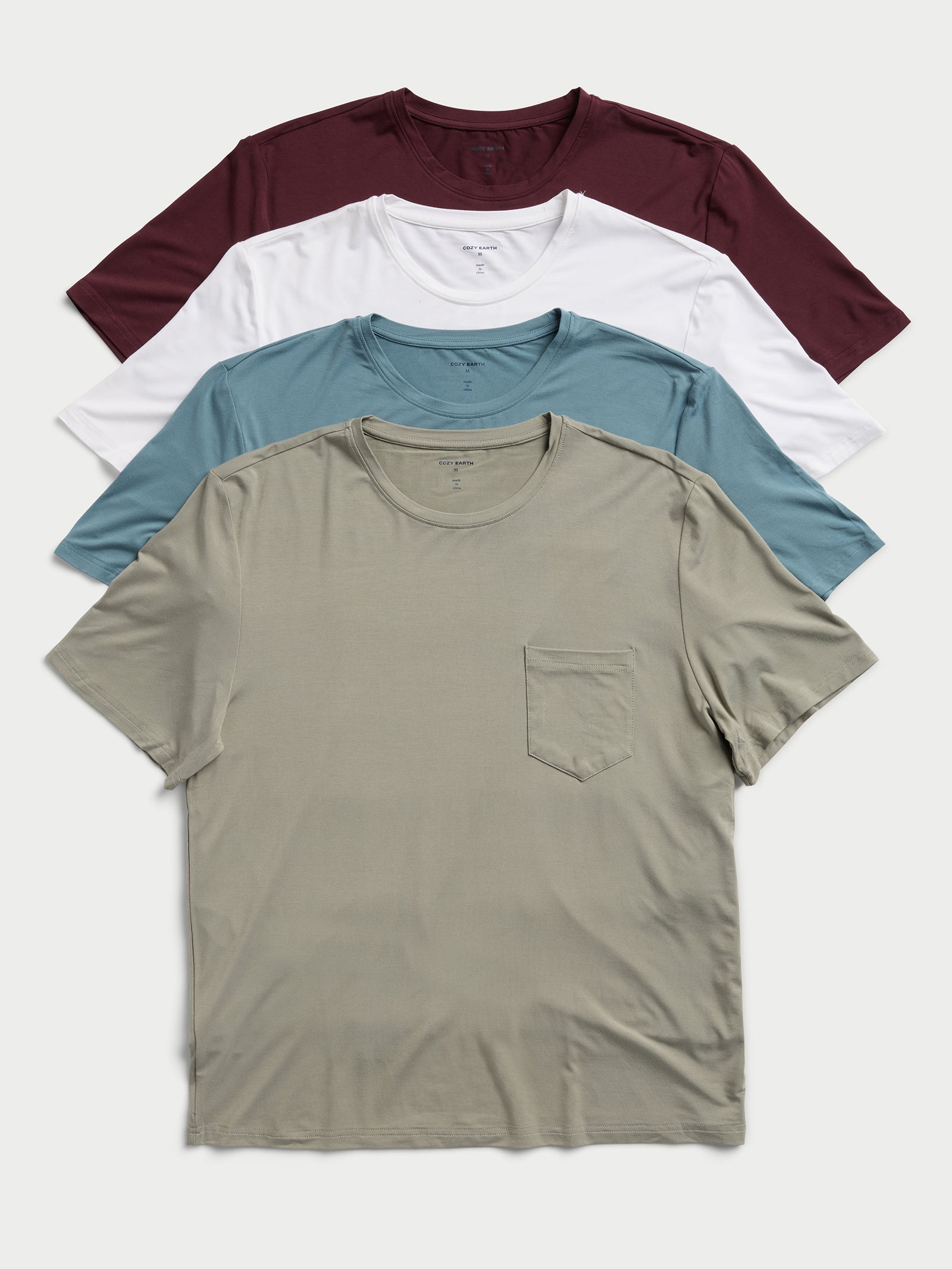 A neatly aligned stack of four Men's Stretch-Knit Bamboo Lounge Tees by Cozy Earth: the front tee in olive green with a pocket, followed by blue-gray, white, and maroon colors. 