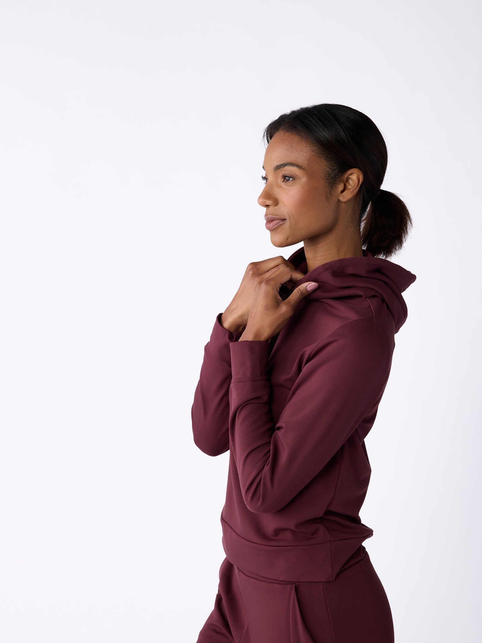 A person wearing the Women's Ultra-Soft Bamboo Hoodie from Cozy Earth stands against a white background, gazing thoughtfully to the side. The maroon hoodie is slightly pulled up near the neck, and the individual has dark hair neatly tied back in a ponytail. 