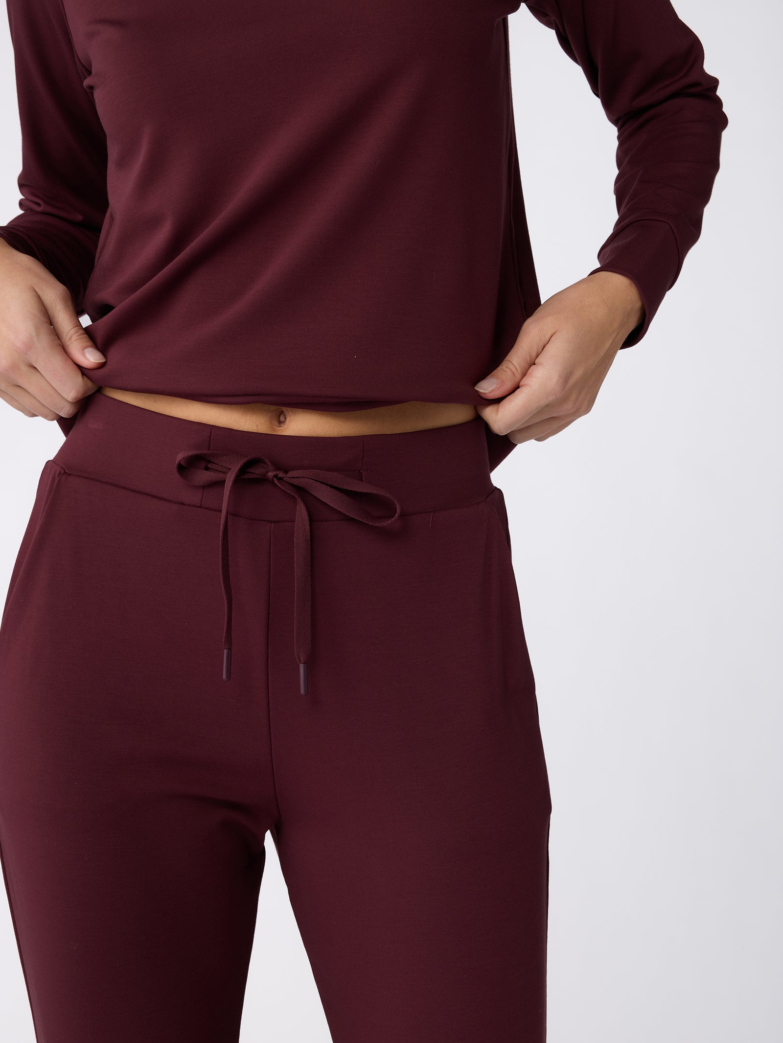 A person dressed in a maroon long-sleeve top and Cozy Earth's Women's Bamboo Jogger Pant with a matching drawstring is adjusting their shirt against a plain white background. 