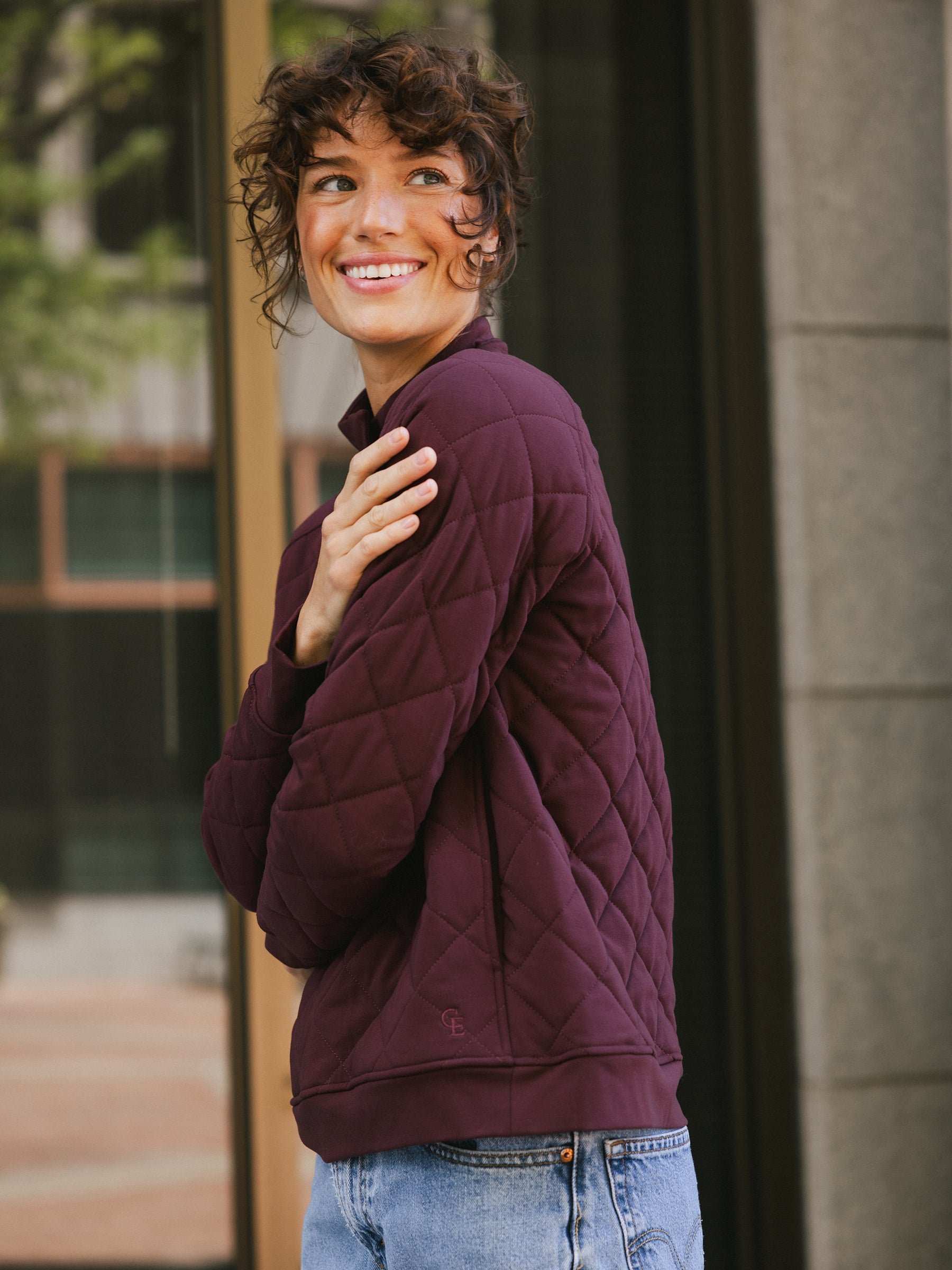 A woman with short curly hair smiles while looking over her shoulder. She is wearing a Cozy Earth Women's Ultra-Soft Bamboo Quilted Snap Pullover in dark purple and light blue jeans. The background features a large window and an exterior wall of a building. 