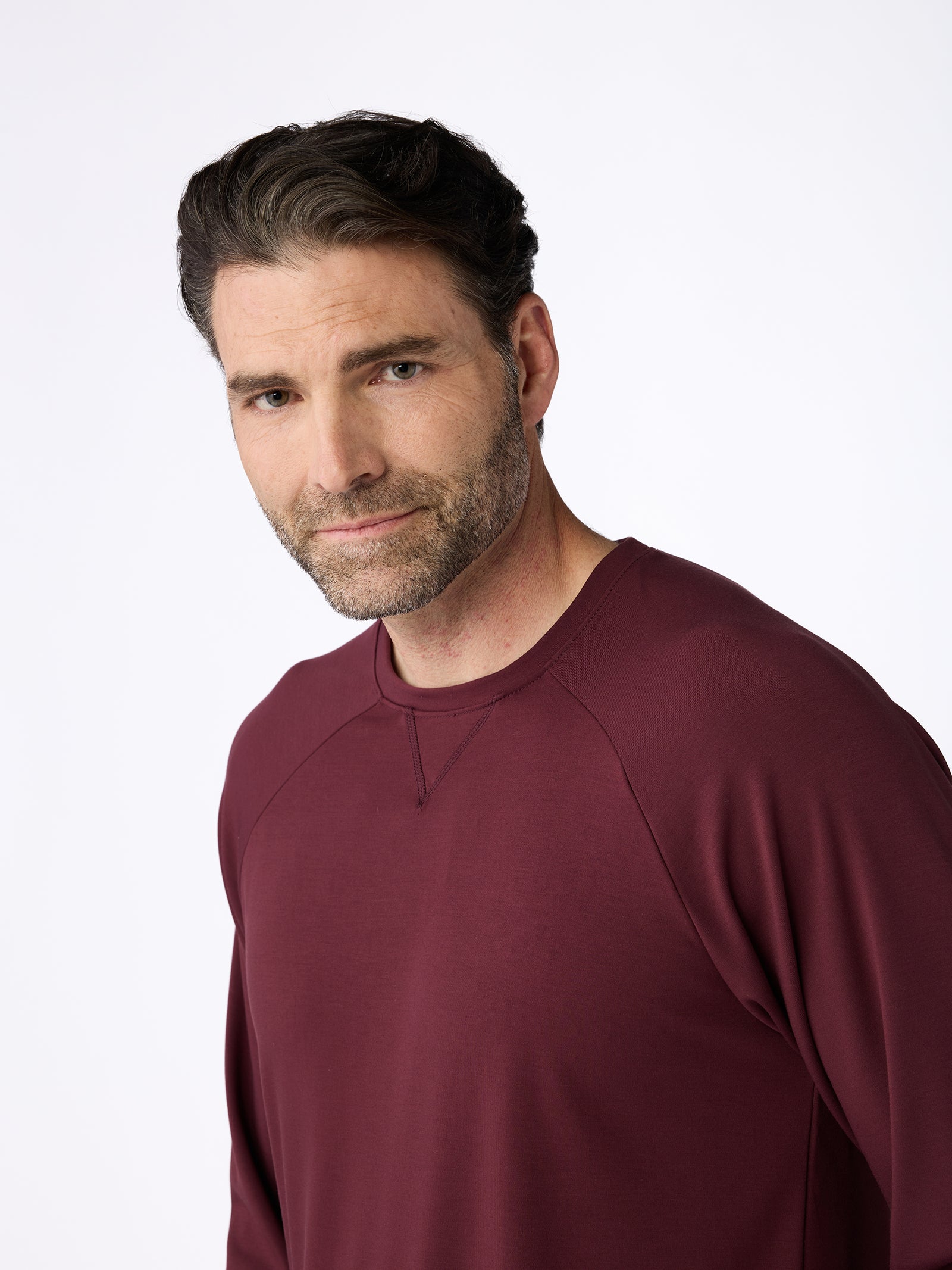 A person with short dark hair and a beard is wearing a maroon Men's Ultra-Soft Bamboo Pullover Crew by Cozy Earth, looking towards the camera with a neutral expression against a plain light background. 