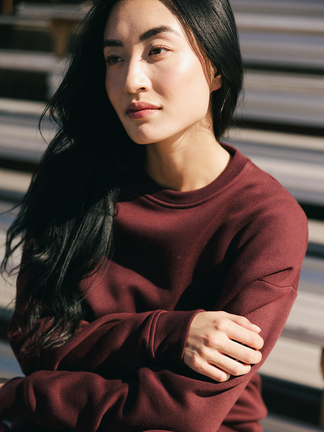 A person with long, dark hair, wearing a maroon Women's CityScape Crewneck by Cozy Earth, is sitting outdoors. Their arms are crossed as they gaze slightly to the side with a calm, contemplative expression. Sunlight illuminates their face, accentuating their features. 