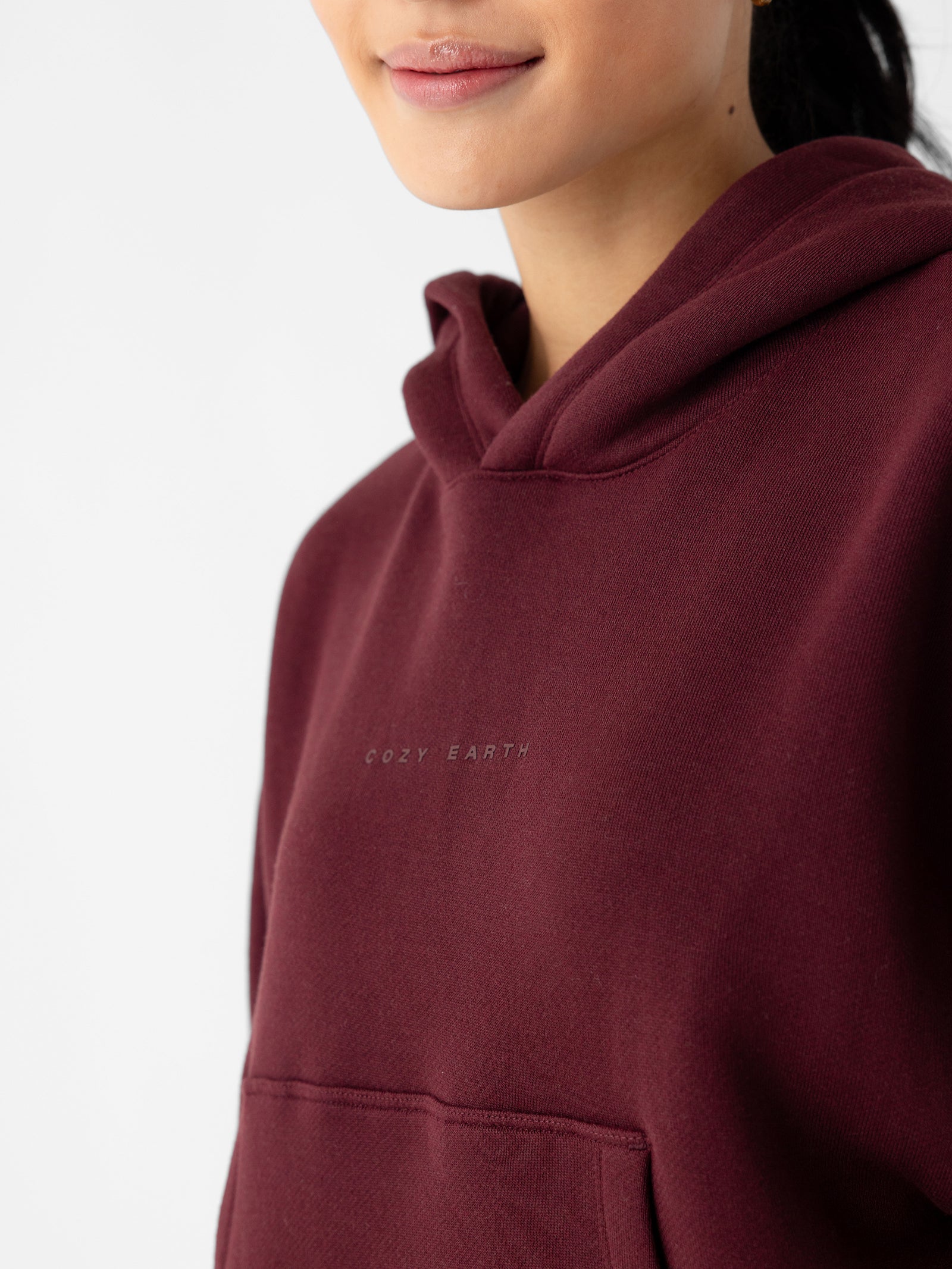 Close-up of a person wearing a Women's CityScape Cropped Hoodie in maroon with the text "Cozy Earth" printed on the chest. Only the lower part of the person's face is visible, along with the upper portion of the hoodie and its front pocket. The background is plain white. 