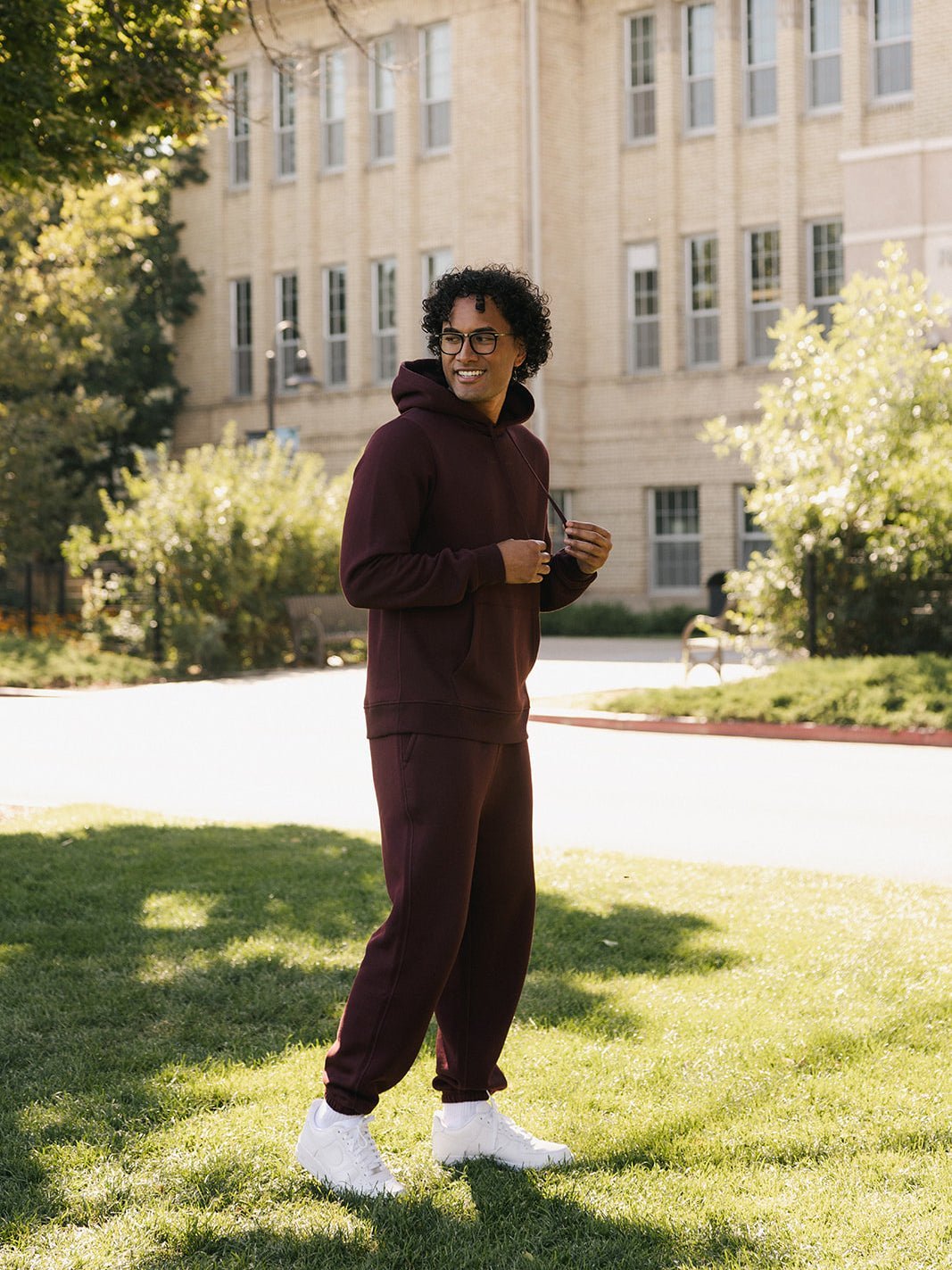 A person with curly hair, wearing glasses, and dressed in a maroon Men's CityScape Hoodie and matching sweatpants from Cozy Earth, stands smiling on a green lawn in front of a beige brick building. Bright sunlight casts shadows, and trees and bushes are visible in the background. 