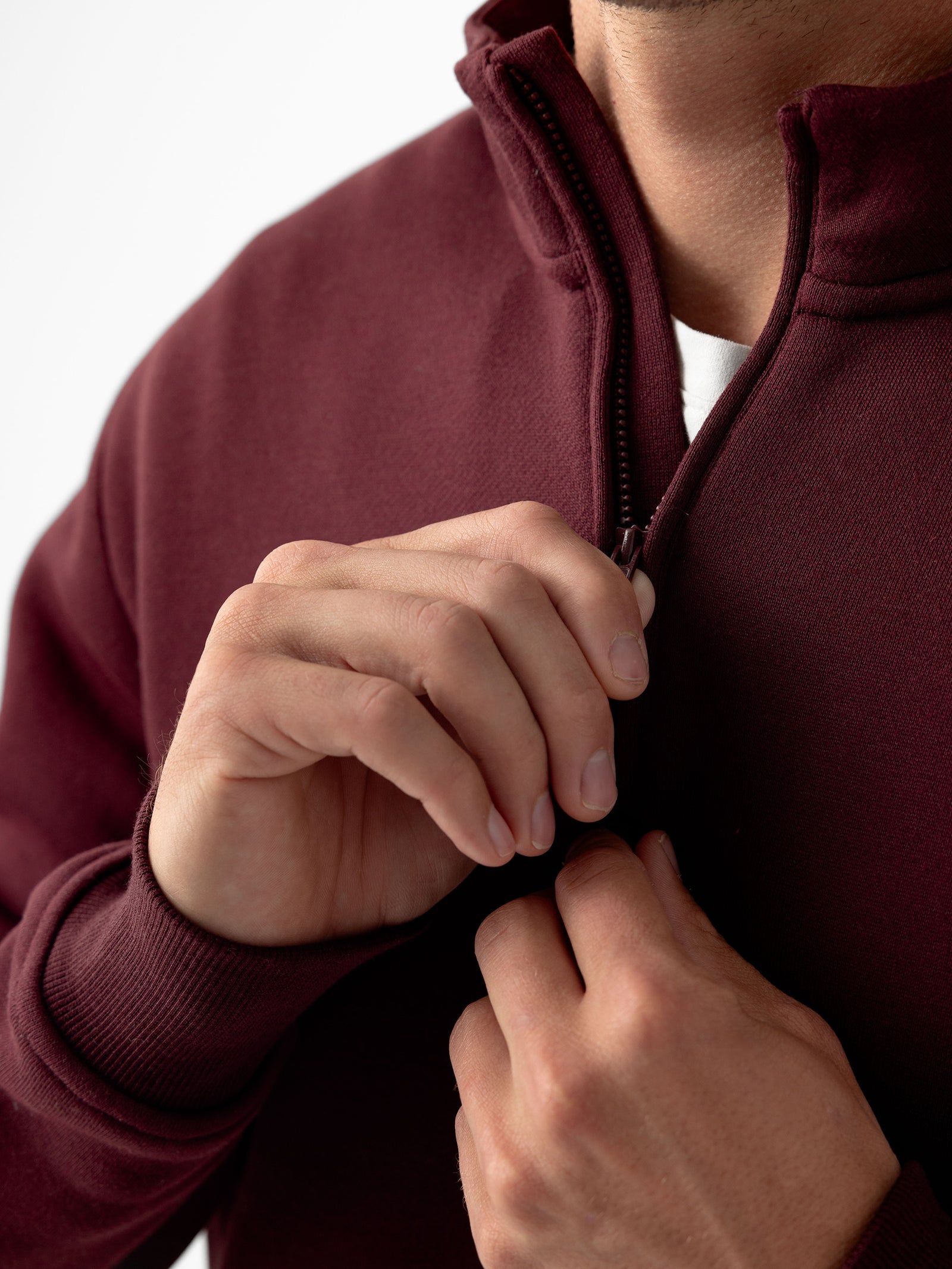 Close-up of a person zipping up a Cozy Earth Men's CityScape Quarter Zip sweater. The person's hands are visible, one holding the zipper pull while the other steadies the maroon fabric. The background is plain white. 