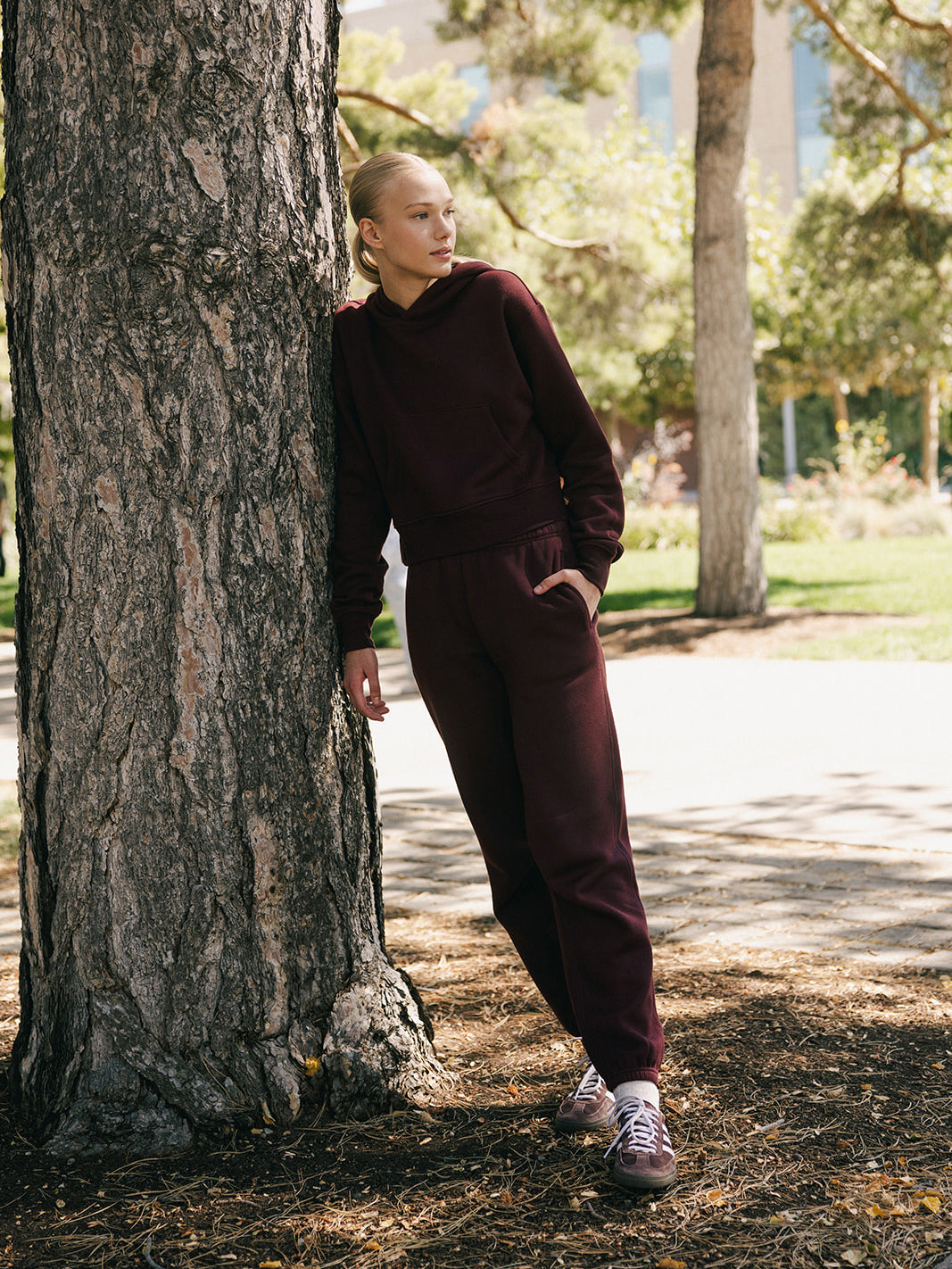 A person with blond hair stands casually against a tree in a park, wearing a matching maroon hoodie and the Women's CityScape Sweatpant by Cozy Earth, along with pink sneakers. The background features greenery and a building, suggesting a serene and sunny day. 