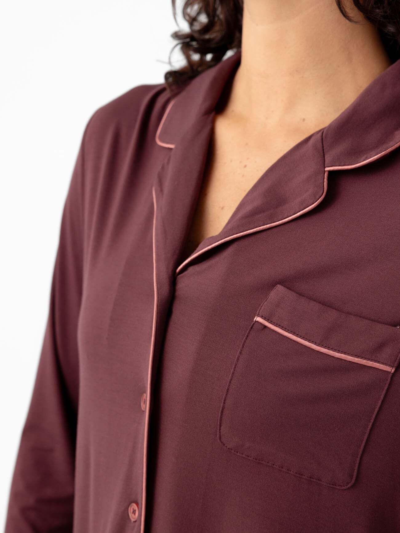 The image features a close-up of someone in a Cozy Earth Last Chance Women's Bamboo Stretch-Knit Long Sleeve Pajama Set top, colored burgundy with pink piping on the collar, pocket, and buttons. The backdrop is plain white. 