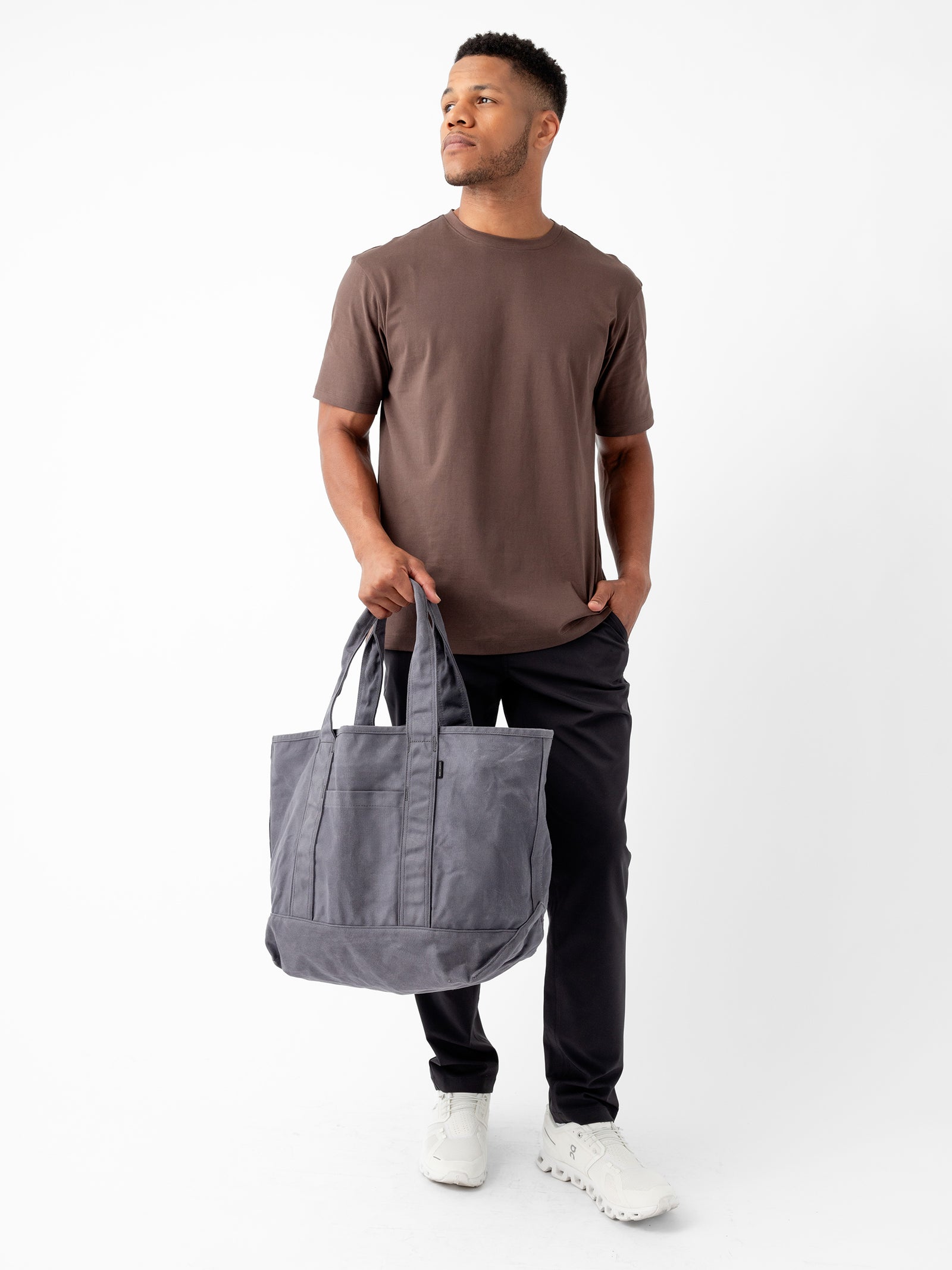 A man stands against a plain white background, wearing Cozy Earth's brown Men's All Day Tee, black pants, and white shoes. He holds a large gray tote bag with his right hand, while his left hand rests in his pocket. He gazes to the side with a calm expression. 