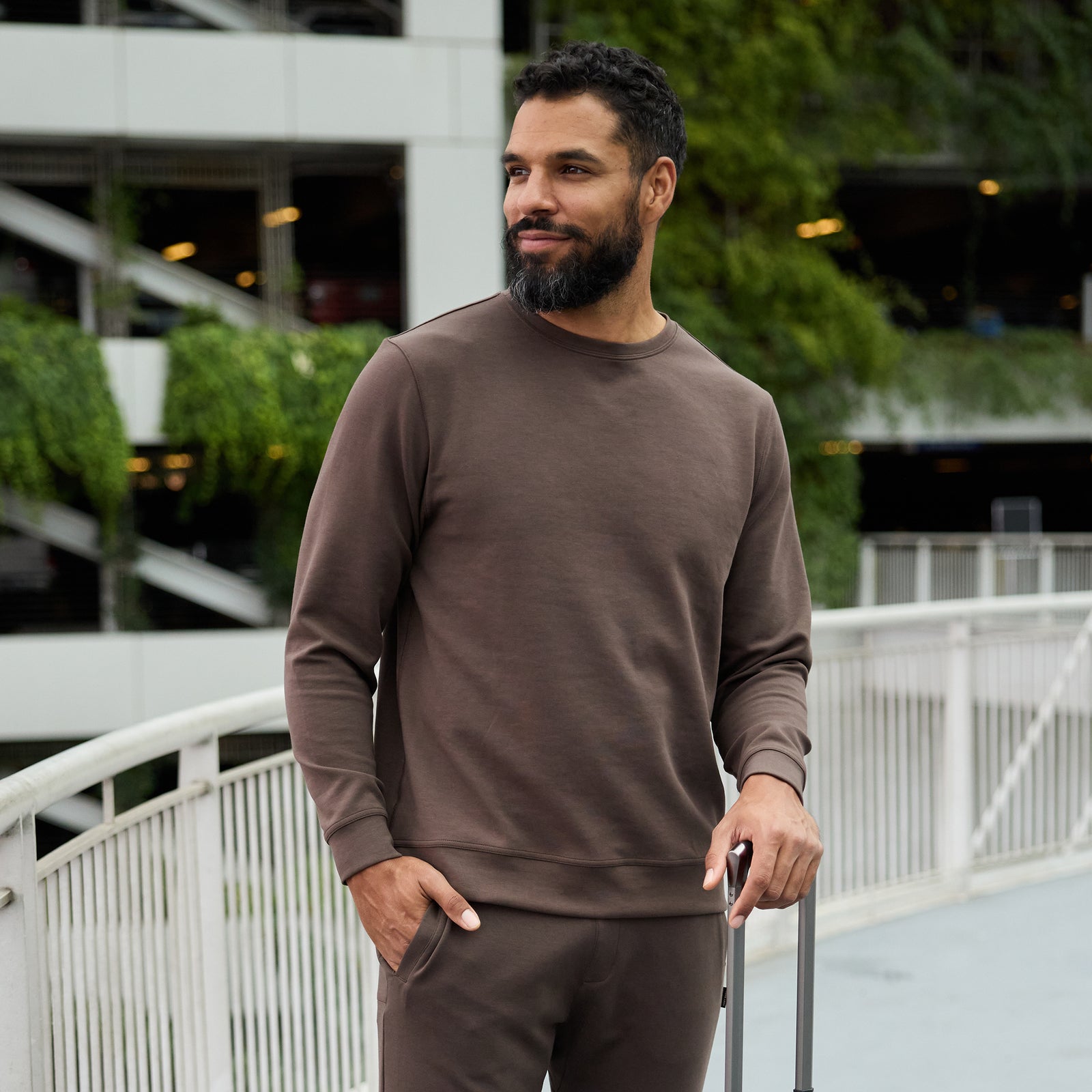 A man with a beard stands outside on a walkway, casually dressed in Cozy Earth's Men's StretchTech Crewneck and matching pants. He is holding the handle of a suitcase and looking off to the side with a slight smile. Lush greenery and part of a multi-storey parking structure are in the background. 