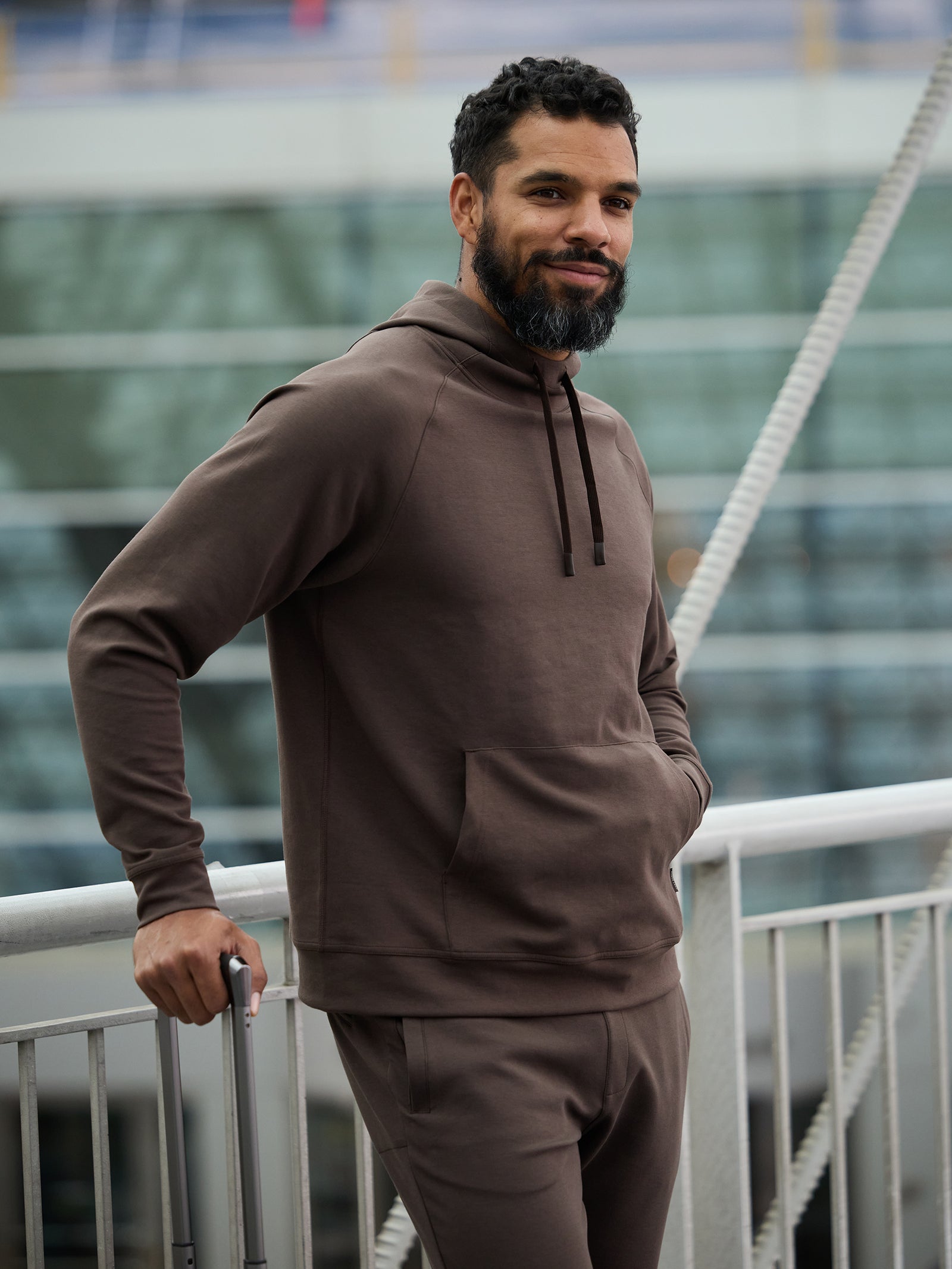 A man with a beard is standing outdoors on a bridge, leaning on the railing with one hand and holding a suitcase handle with the other. He is dressed in Cozy Earth’s Men's StretchTech Hoodie and matching sweatpants, smiling slightly and looking at the camera. 