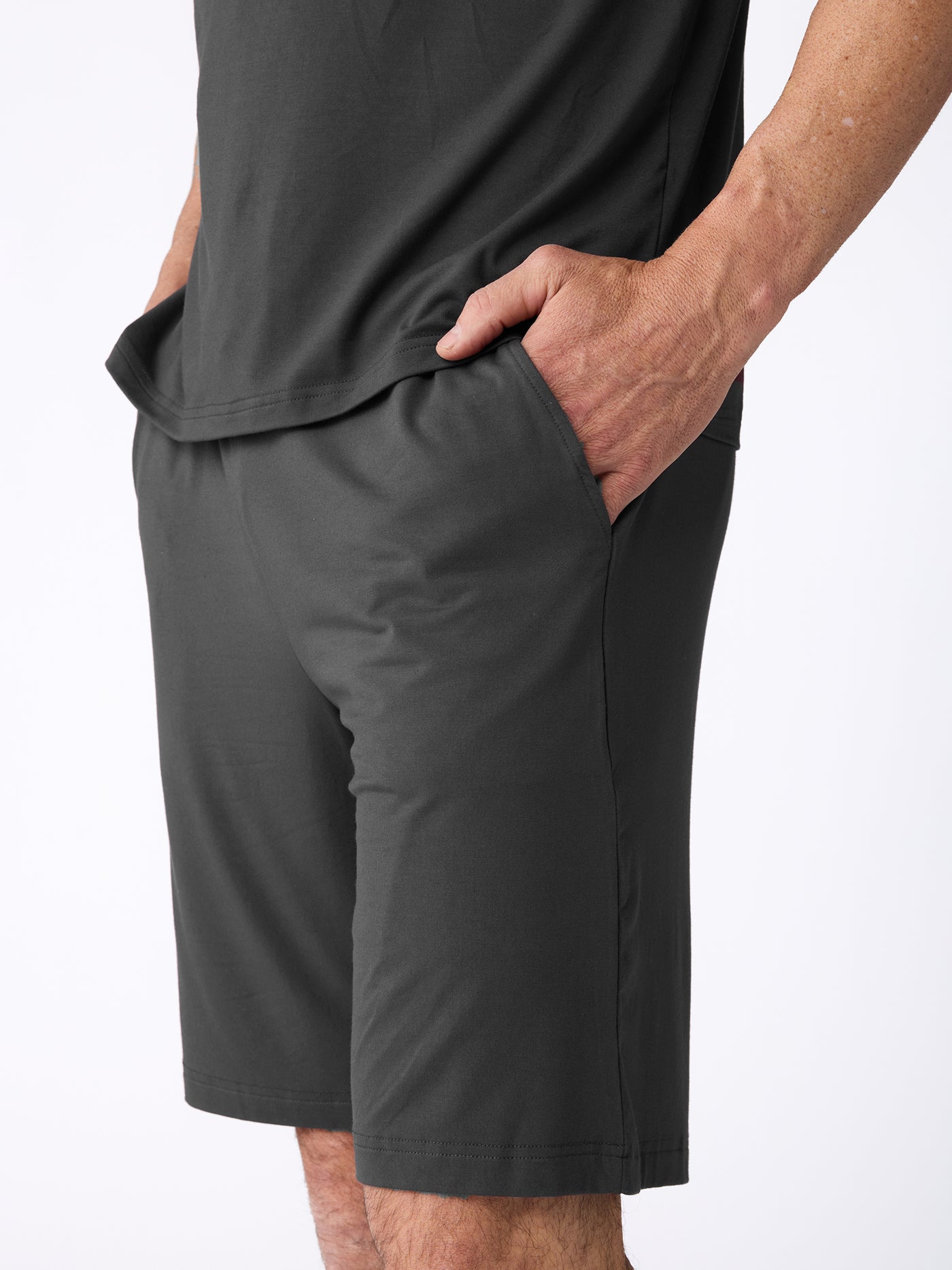 A man models the Cozy Earth Men's Bamboo Stretch-Knit Short Sleeve Pajama Set in dark gray, standing against a plain white background with one hand in his shorts pocket. 