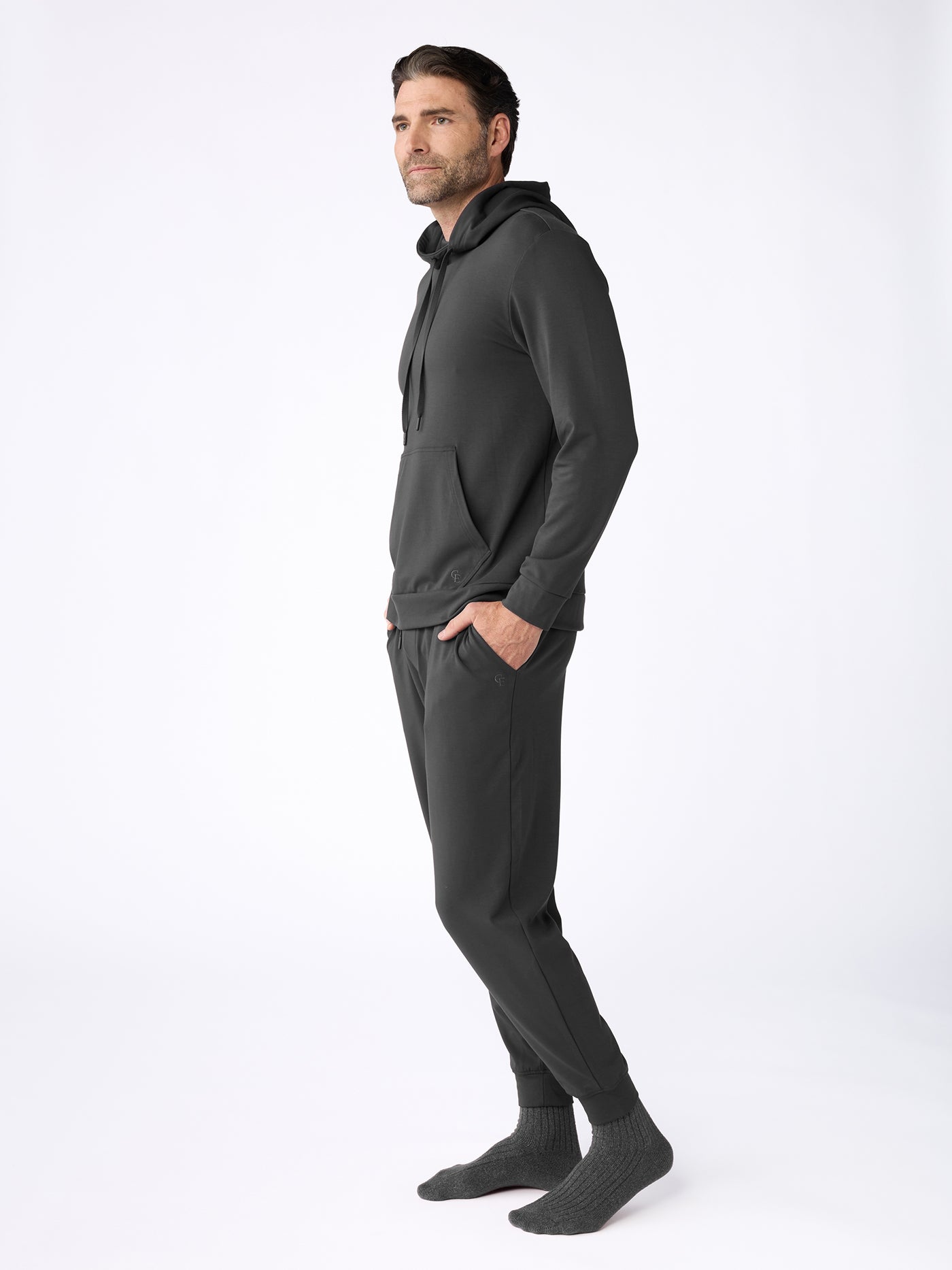 A man with short brown hair and a beard stands sideways against a white background, wearing the Cozy Earth Men's Brushed Bamboo Hoodie & Jogger Set in dark gray with gray socks. 