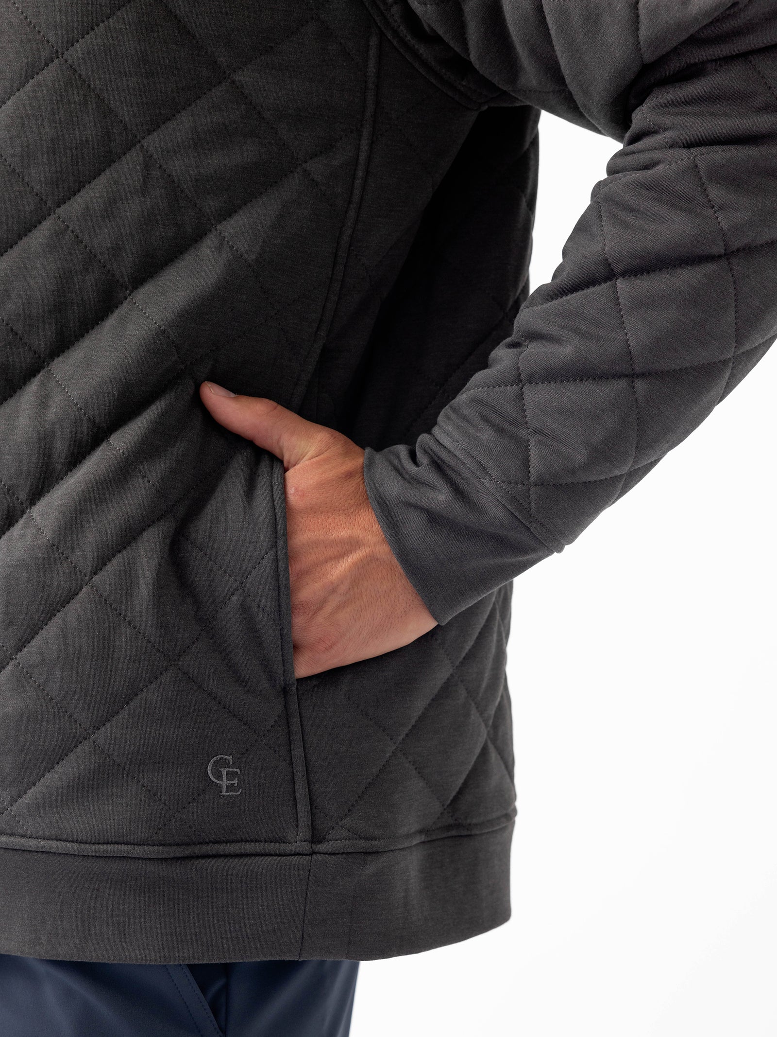 Close-up of a person wearing the Cozy Earth Men's Ultra-Soft Bamboo Quilted Snap Pullover in dark gray, with a hand resting in a side pocket. The sleeve features diagonal stitching, and the bottom of the pullover has a small logo with the letters "GE." The person is also wearing dark blue pants. 