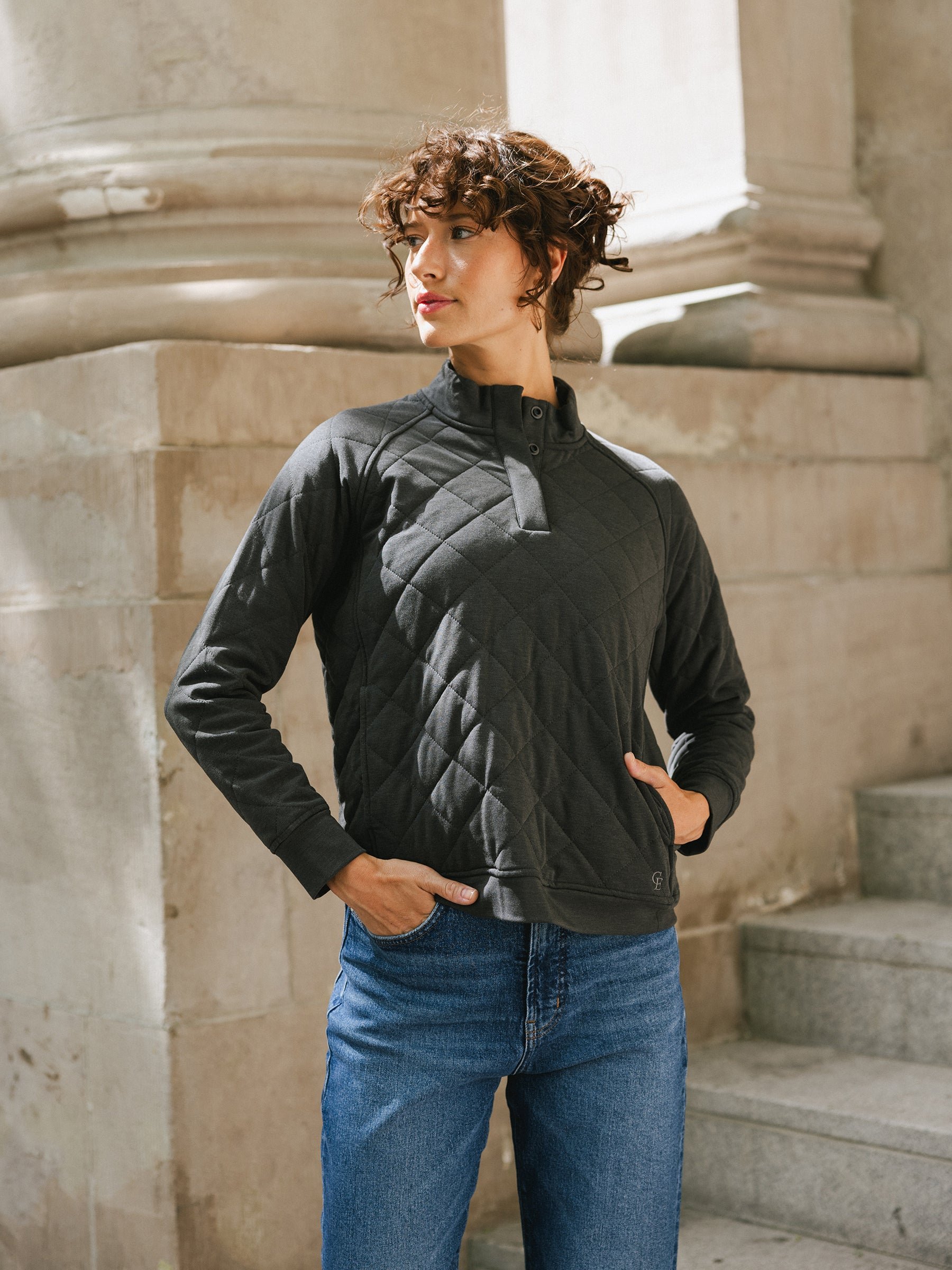 A person with short curly hair stands outdoors near stone columns and stairs. They are wearing a Cozy Earth Women's Ultra-Soft Bamboo Quilted Snap Pullover and blue jeans, with one hand in their pocket and the other resting on their hip, gazing thoughtfully to the side. 