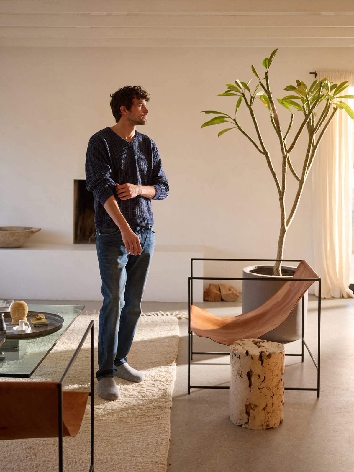 A person stands in a minimalist living room featuring a large plant, a unique wooden chair, and contemporary furniture. Dressed in jeans and a dark shirt by Cozy Earth, they gaze to the side in the well-lit space of neutral tones. 