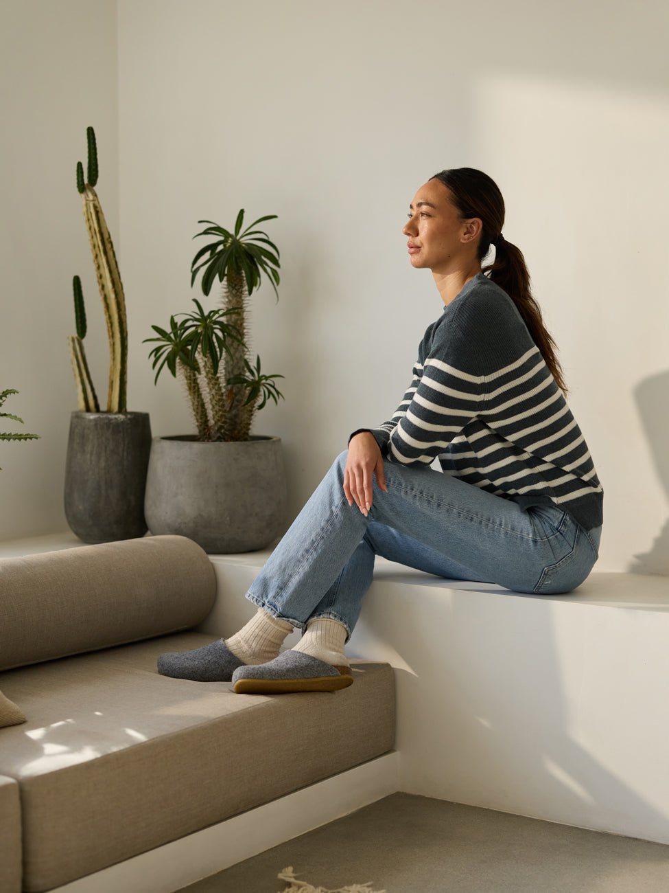 A person wearing a striped sweater and jeans sits on a ledge in a sunlit room, looking to the side with a relaxed expression. They are wearing Lakehouse Clogs by Cozy Earth. Potted plants, including a cactus, decorate the floor in the background. 