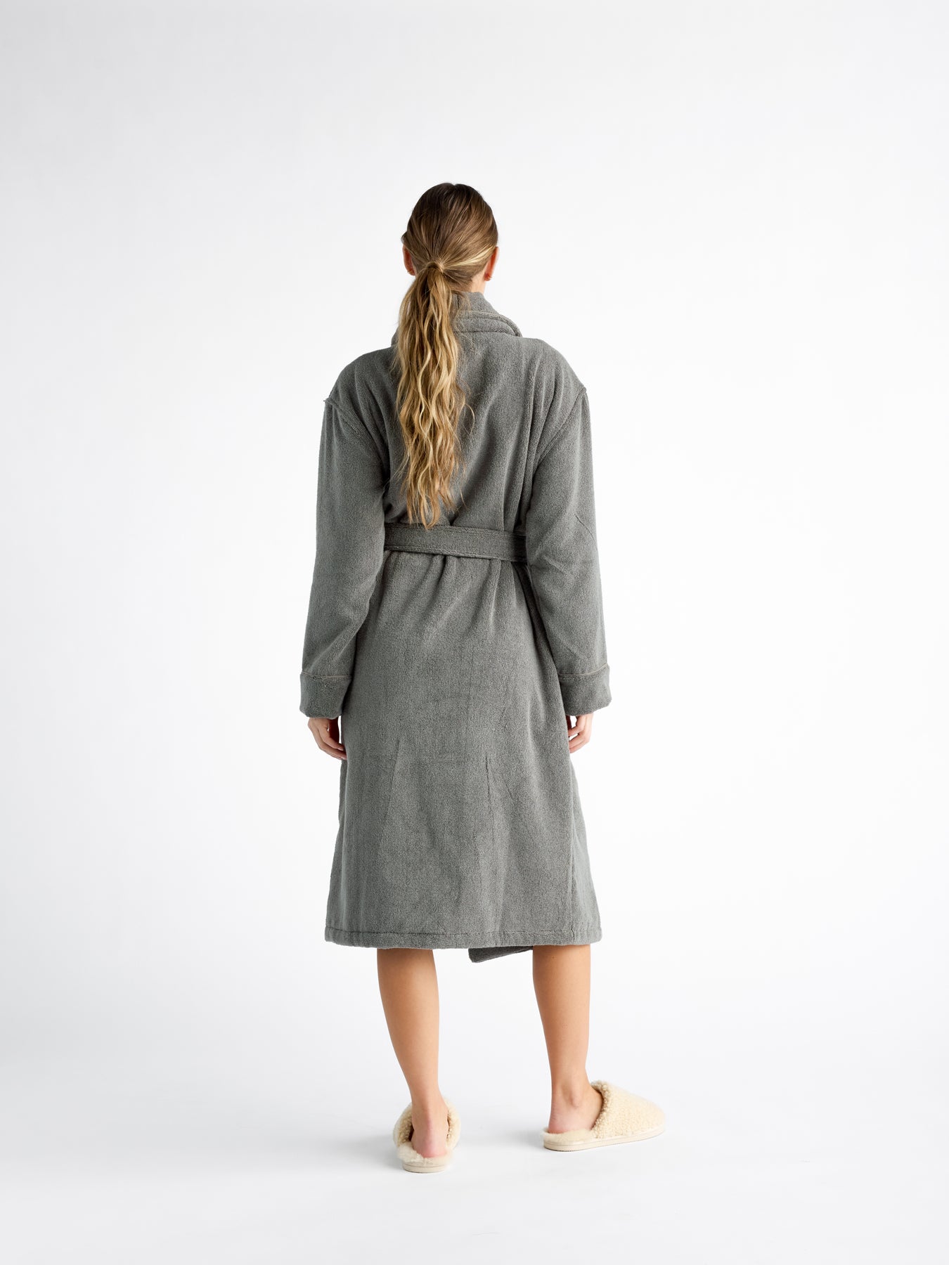 A woman is turned away, wearing a Luxe Bath Robe by Cozy Earth in grey. The robe is belted at the waist, and the person's long, blonde hair hangs down their back against a plain white background. 