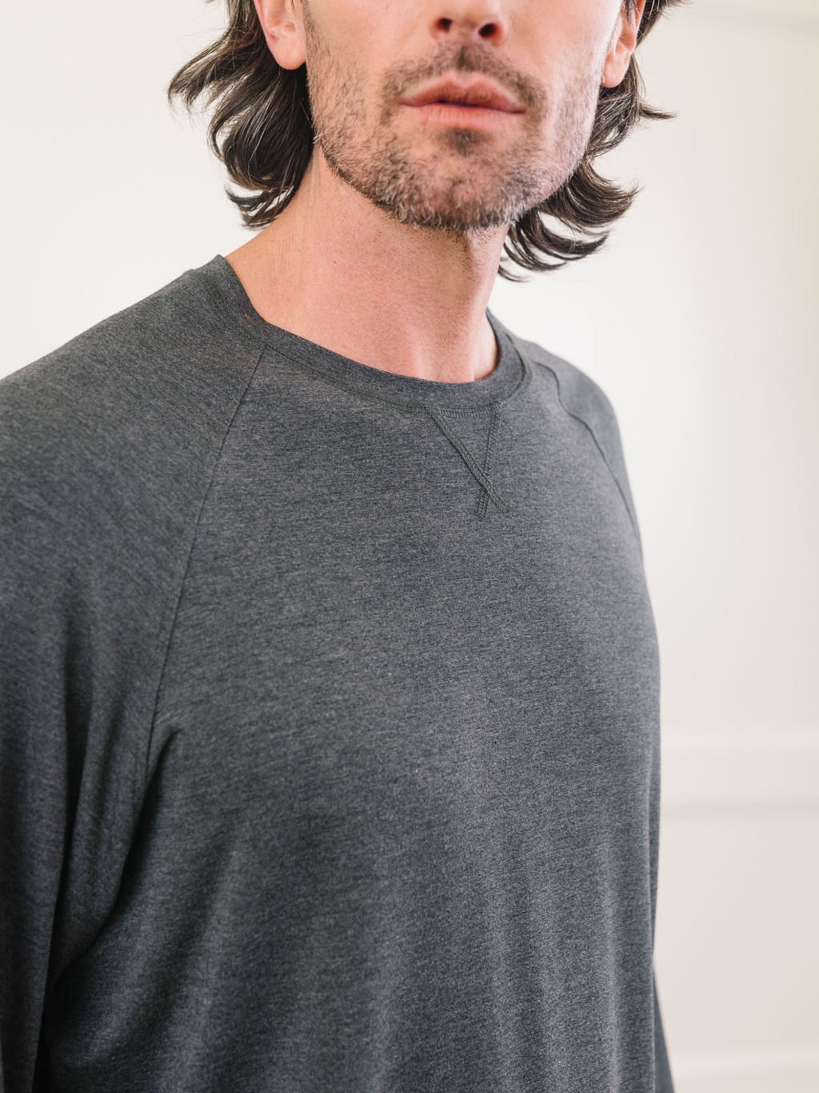 A man with dark shoulder-length hair and slight facial hair is wearing a long-sleeve, dark gray that appears to be part of the Men's Stretch-Knit Long Sleeve Bamboo Pajama Set by Cozy Earth. The photo is cropped to show his upper torso, with his head slightly tilted to the left. The background is out of focus. 