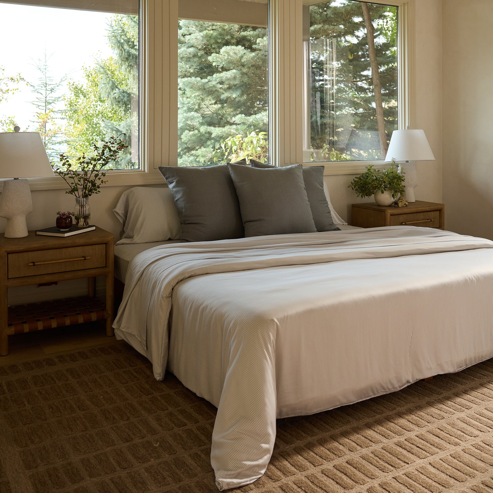 A cozy bedroom showcases a neatly made bed featuring Bamboo Pillowcases from Cozy Earth, with white bedding and gray accent pillows. Two wooden nightstands with white lamps and plants flank the bed, while large windows offer a view of lush greenery outside, filling the room with natural light. 