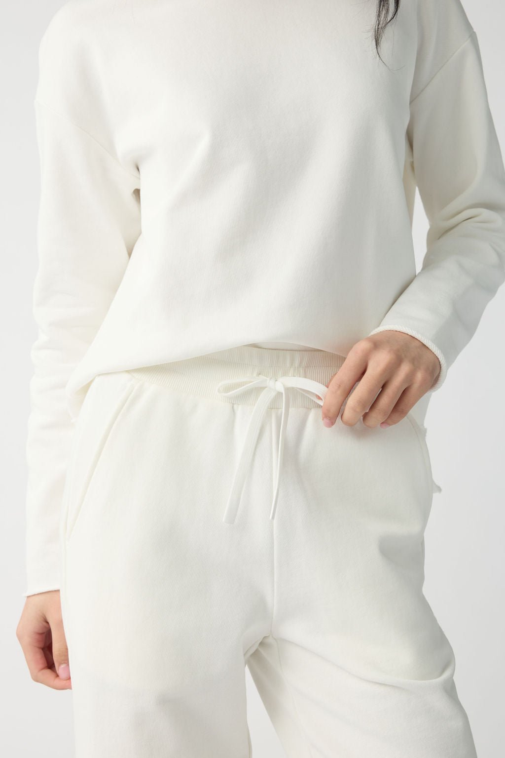 A person is wearing Cozy Earth's Women's Chelsea Set, featuring a white long-sleeve sweatshirt and drawstring sweatpants. They adjust the drawstring in an all-white setting. 