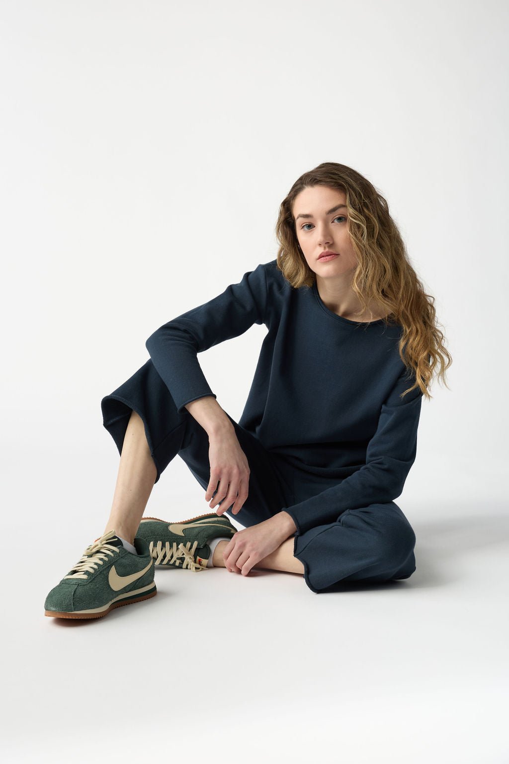 A person with long wavy hair sits on the floor in a relaxed pose. They wear the Women's Chelsea Set by Cozy Earth, featuring a dark blue long-sleeve outfit, paired with green sneakers against a plain white background. 