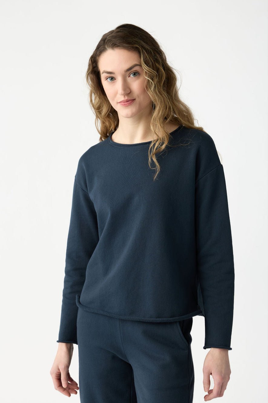 A person with long, wavy hair is wearing the Eclipse Women's Chelsea Pullover and matching pants from Cozy Earth, standing against a plain white background and looking directly at the camera with a neutral expression.