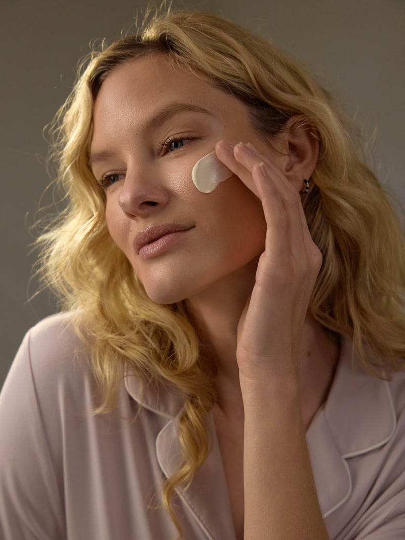 With long, wavy blonde hair, a person applies Cozy Earth's Clay Mask to their cheek, appearing at ease. They're dressed in a pale pink top with a gentle smile. The neutral background emphasizes their skincare ritual.