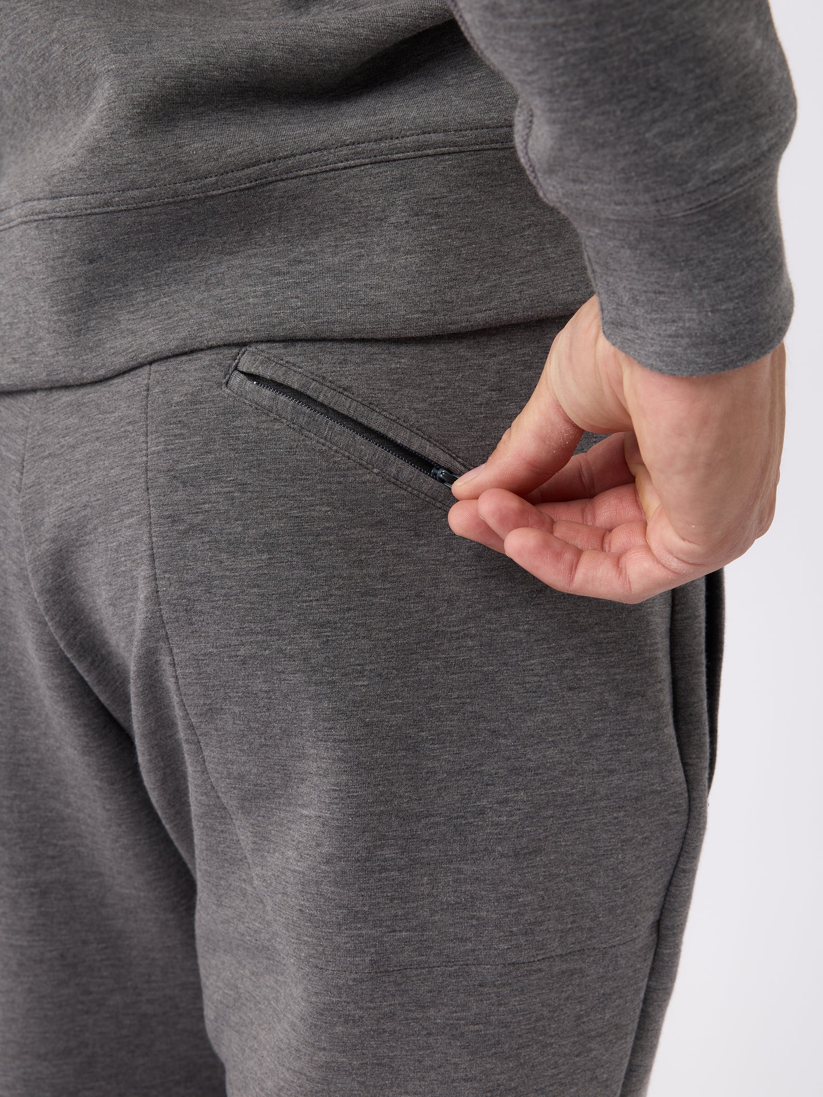 A person wearing the Men's StretchTech Jogger by Cozy Earth is shown from behind, with their hand reaching to open a zipper on the back pocket. The focus is on the pocket and the hand. 