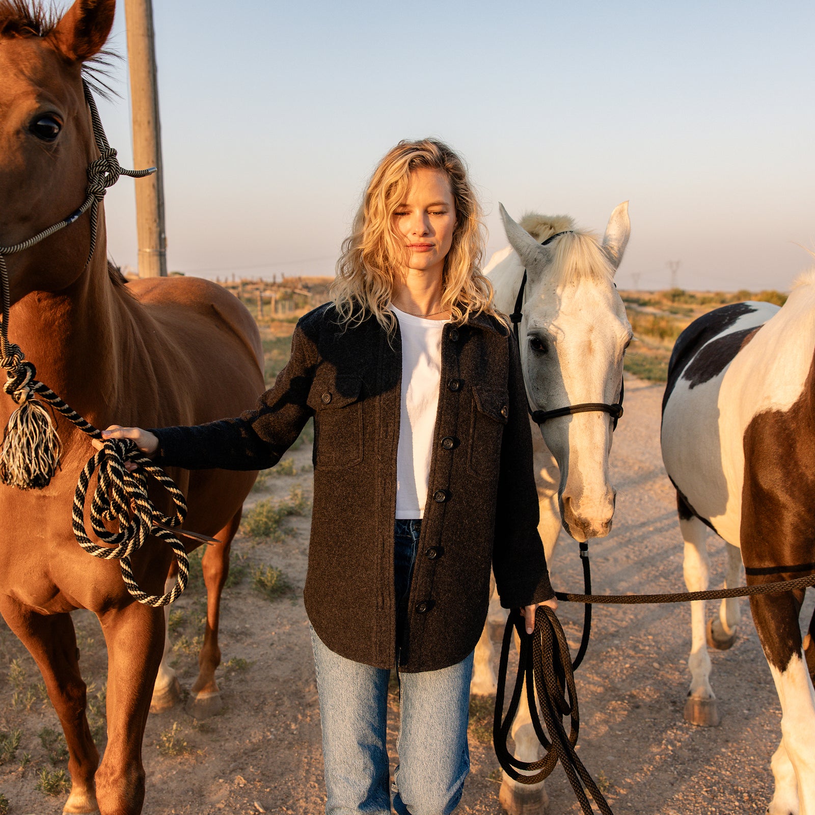 A woman with long, curly blonde hair stands outdoors holding the reins of three horses. She is wearing a Cozy Earth Women's Boucle Shacket over a white shirt and blue jeans. The sun is low on the horizon, casting a warm glow over the scene. The background includes a dirt path and a fence. 