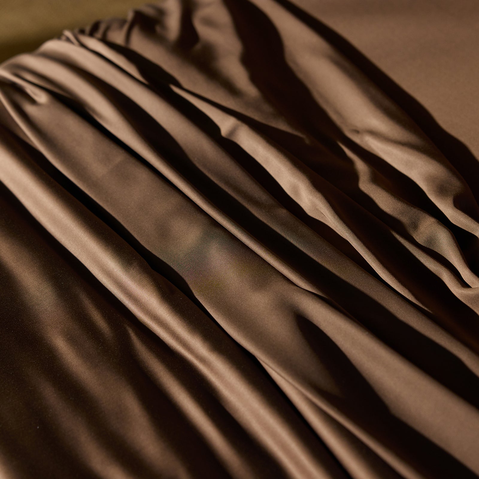 A close-up of the Bamboo Duvet Cover by Cozy Earth, showcasing its smooth, brown satin fabric draped in gentle folds. The texture of the material reflects light, creating a soft sheen and highlighting the fluid lines of the cover's arrangement. 