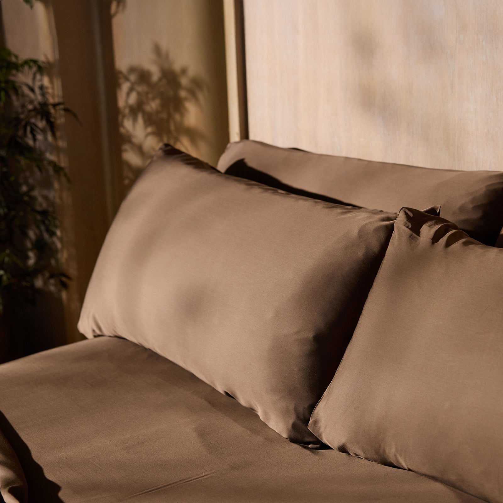 A neatly made bed showcases the Bamboo Fitted Sheet by Cozy Earth, adorned with brown pillows and bedsheets. The headboard and wall feature a light wooden texture, while dappled shadows of plant foliage hint at nearby greenery. The scene exudes warmth and invitation. 