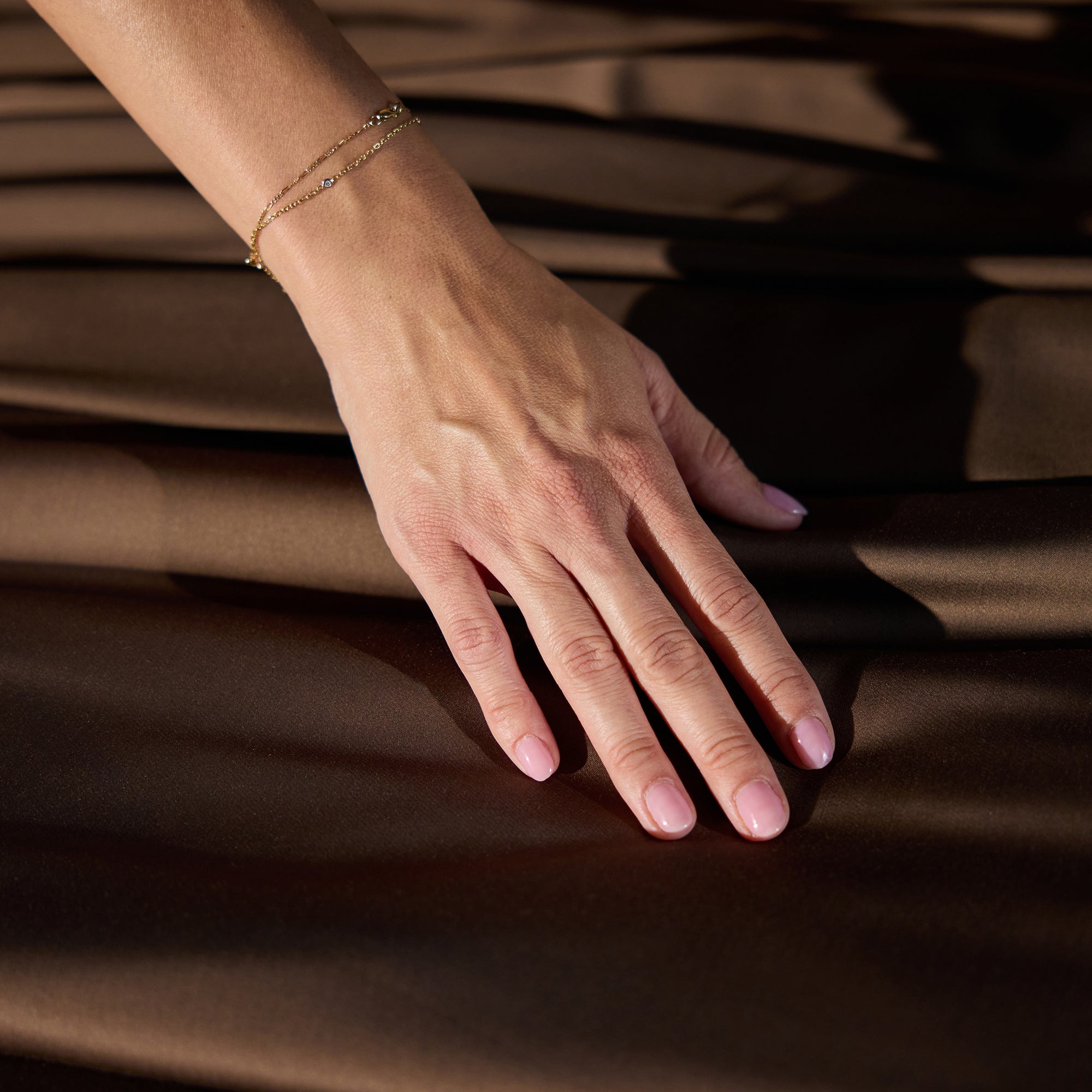 A close-up of a woman's hand resting on smooth, dark brown fabric from the Cozy Earth Bamboo Sheet Set. She is wearing a delicate gold bracelet and has neatly manicured nails with light pink nail polish. The fabric has a soft sheen, creating subtle light and dark contrasts. 