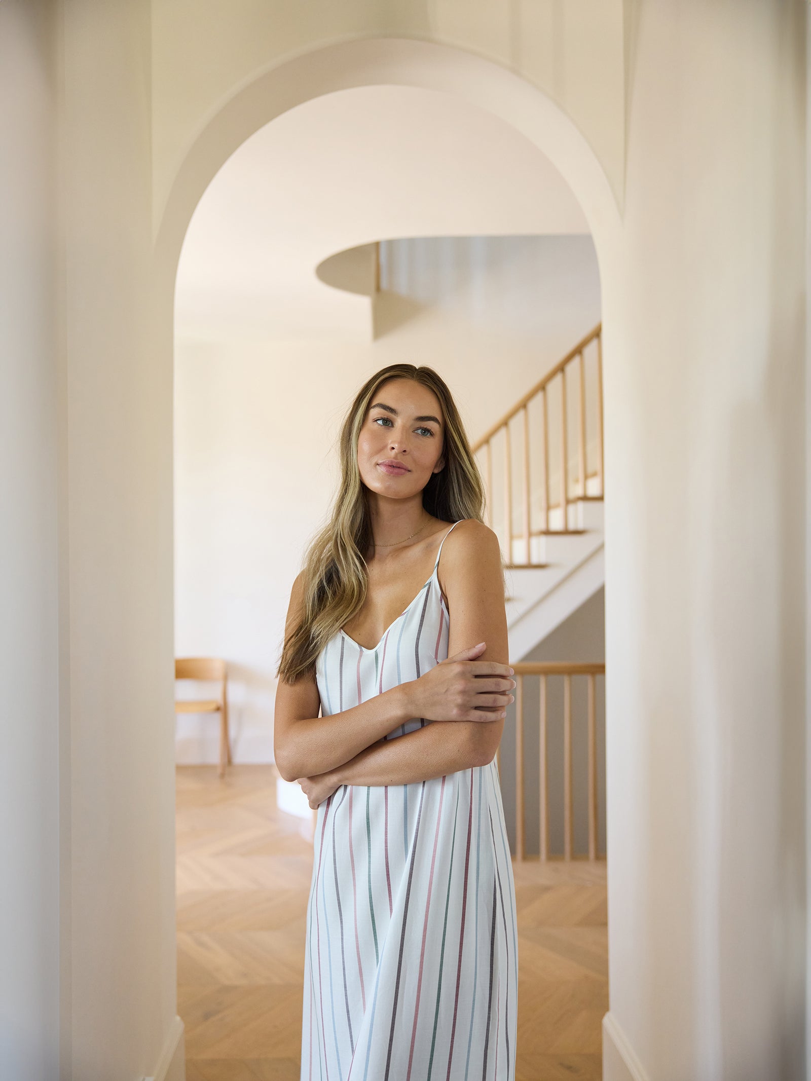 A woman with long hair stands in a bright hallway, dressed in a sleeveless nightgown designed by Cozy Earth. She crosses her arms lightly in front of her body as a staircase and a wooden chair appear softly lit in the background. 