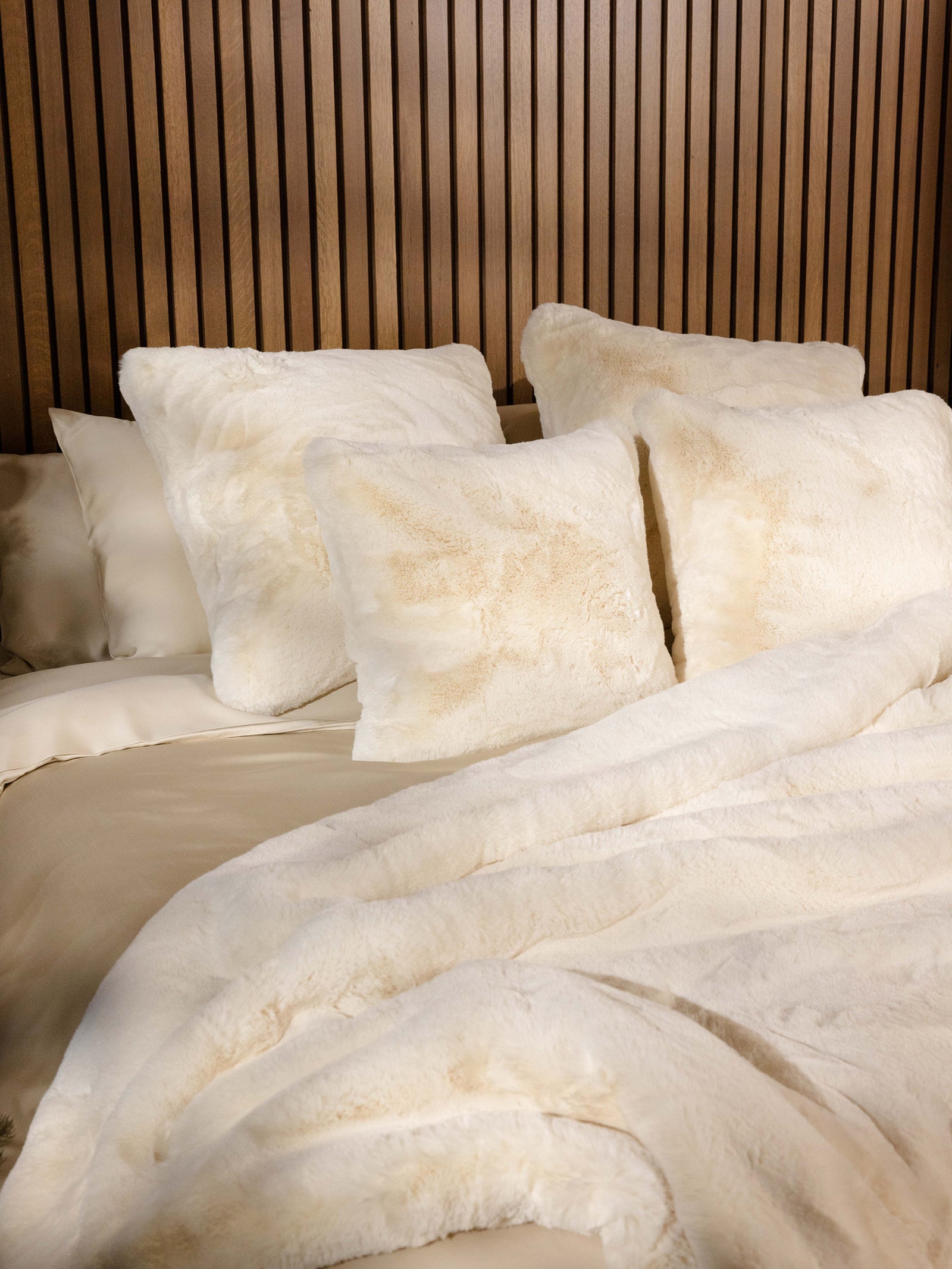 A comfortable bed showcases the Cuddle Pillow from Cozy Earth, featuring fluffy cream color, paired with a matching soft blanket. The setting is enriched by a backdrop of dark wooden paneled walls, creating an inviting and warm atmosphere. 