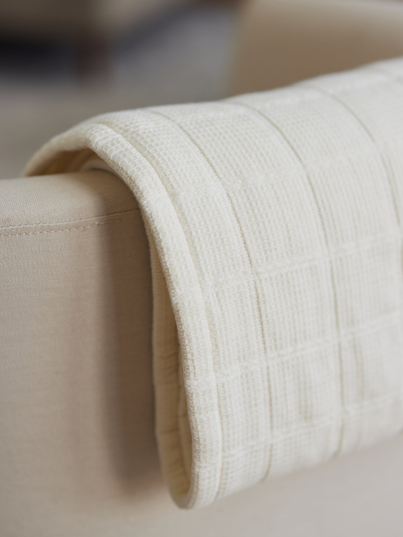 A *cream-colored, Waffle Windowpane Blanket* from Cozy Earth is delicately draped over the armrest of a beige sofa. This knitted throw blanket features a textured grid pattern, enhancing the cozy and warm aesthetic of the furniture piece. The background is softly blurred, keeping the focus on the blanket and sofa. 