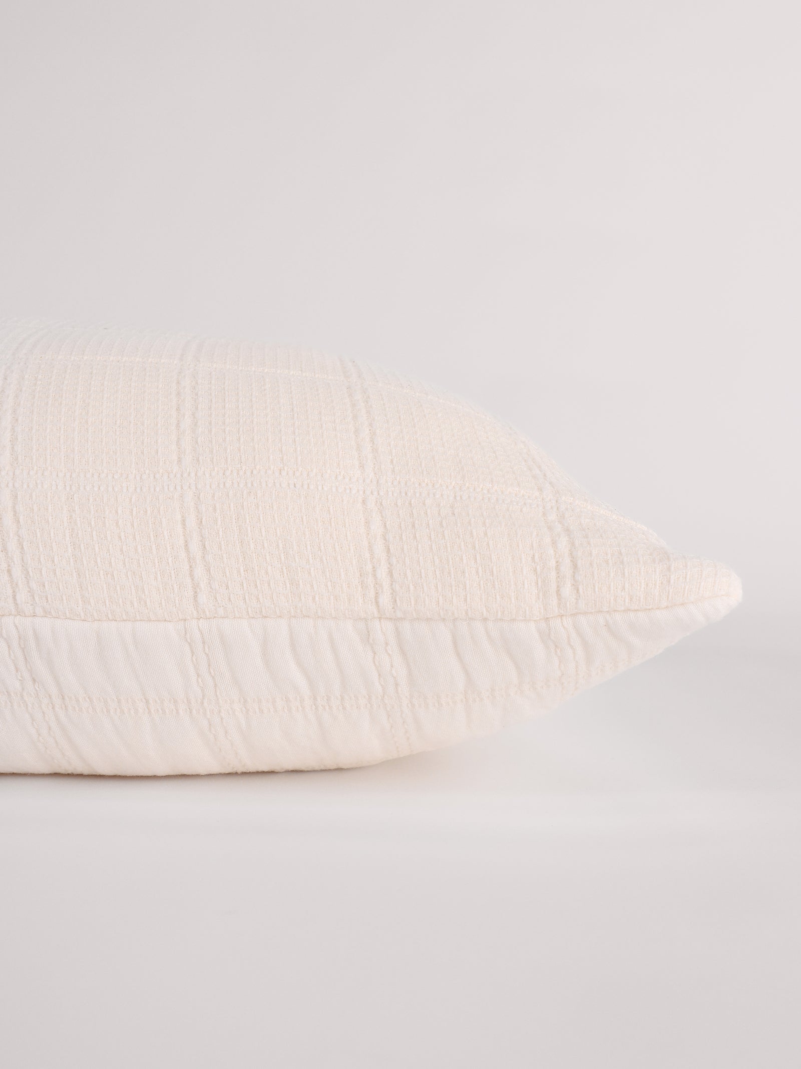 A close-up side view of the Waffle Windowpane Pillow by Cozy Earth, featuring a textured, ribbed cream-colored fabric. The pillow is resting on a white surface against a plain, light gray background. 