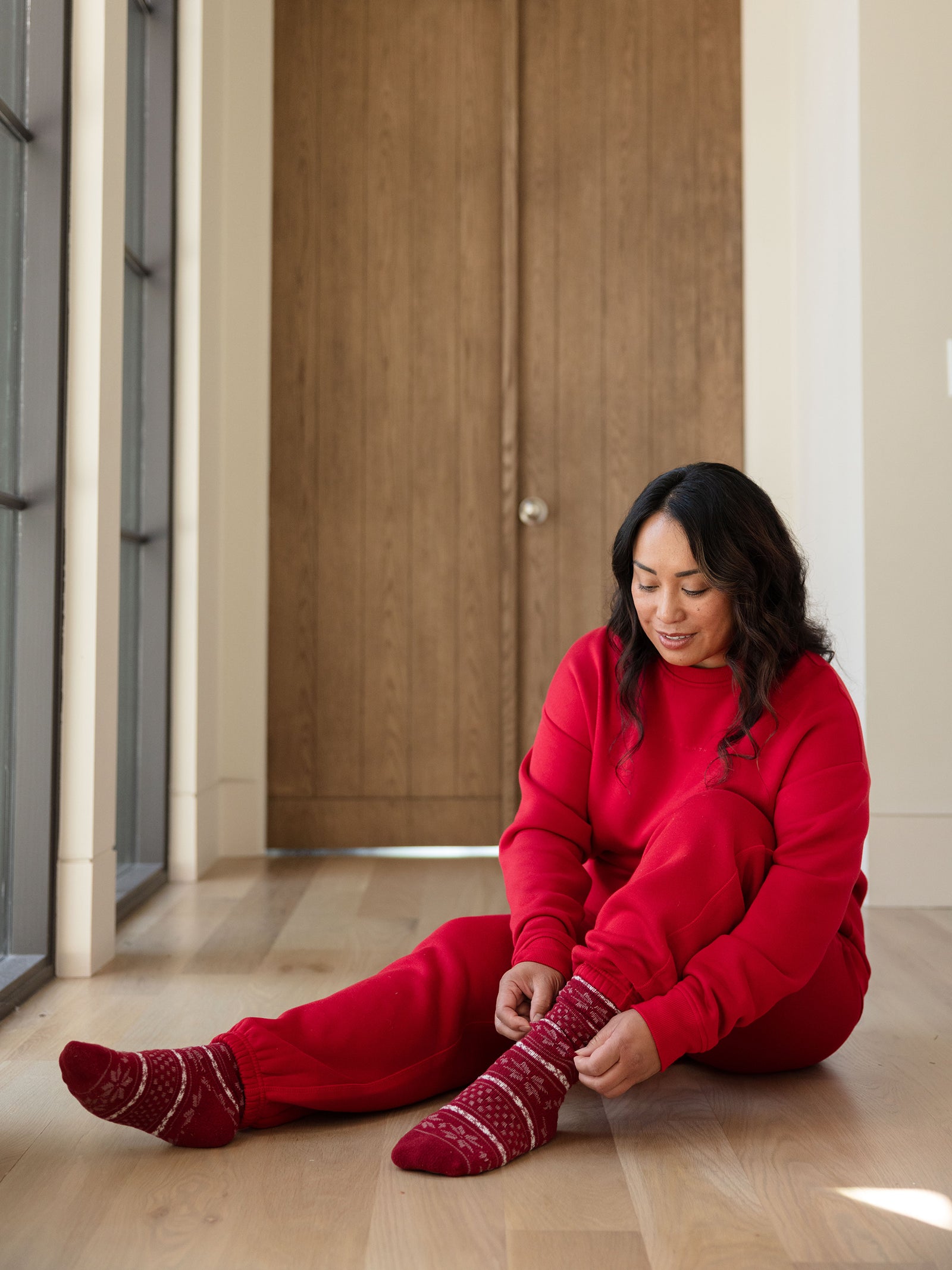 Dressed warmly in a red Women's CityScape Crewneck by Cozy Earth, an individual sits on a light wooden floor, adjusting their red patterned socks. Large wooden doors and tall windows in the background allow natural light to stream in. 