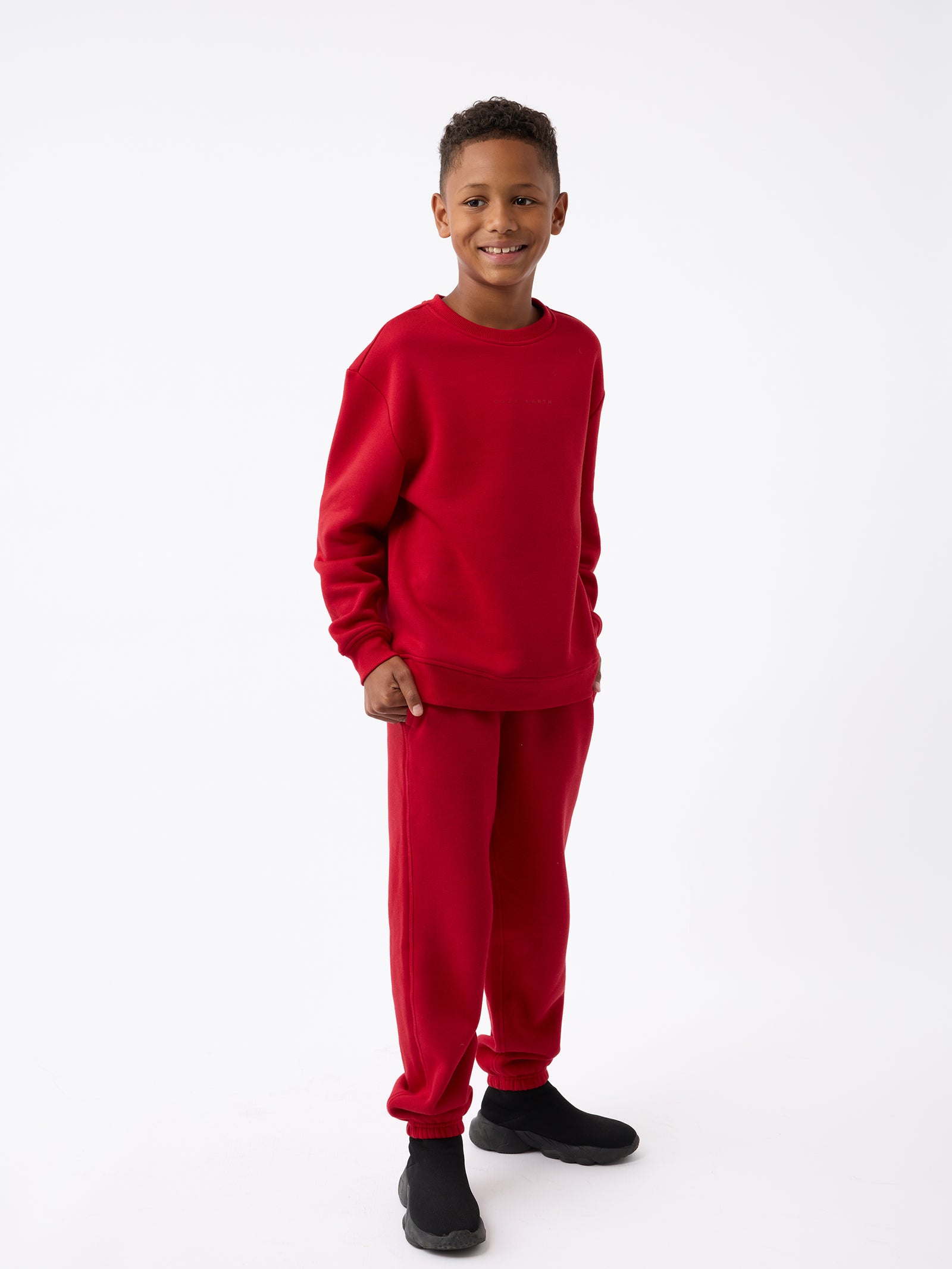 A boy wearing the Kid's CityScape Crewneck by Cozy Earth and black shoes stands smiling against a plain white background. 