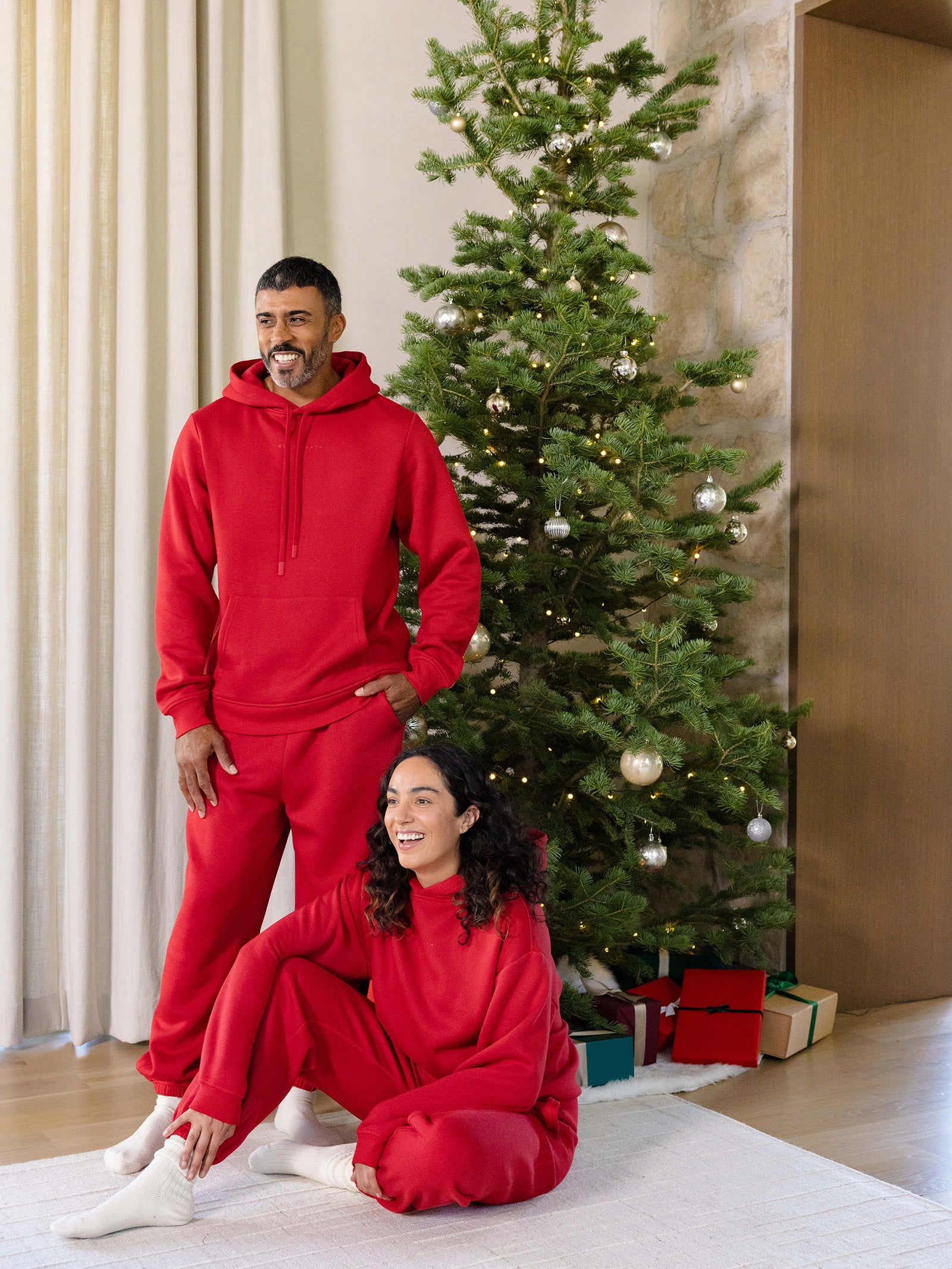 A man and woman in matching outfits pose by a decorated Christmas tree; she is sitting on the floor smiling while wearing the Women's CityScape Hoodie from Cozy Earth, as he stands with hands in pockets. Wrapped gifts are placed under the tree, illuminating the warmly lit room. 