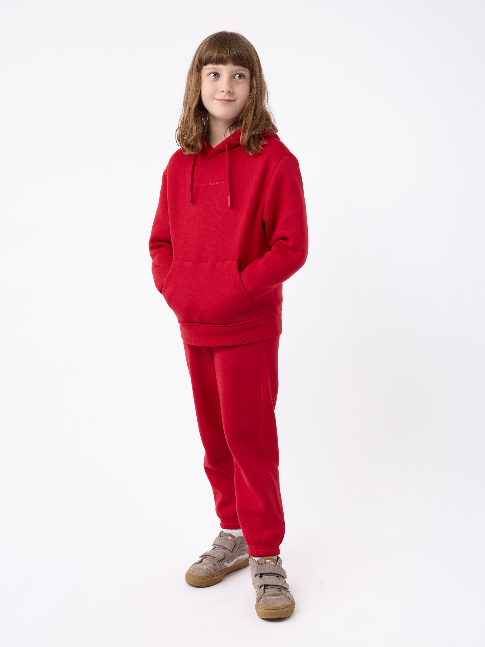 A young person stands against a white background, wearing the Cozy Earth Kid's CityScape Hoodie and matching red sweatpants, their hands tucked into the hoodie pocket as they gaze to the side with a neutral expression. They complete their look with brown shoes. 