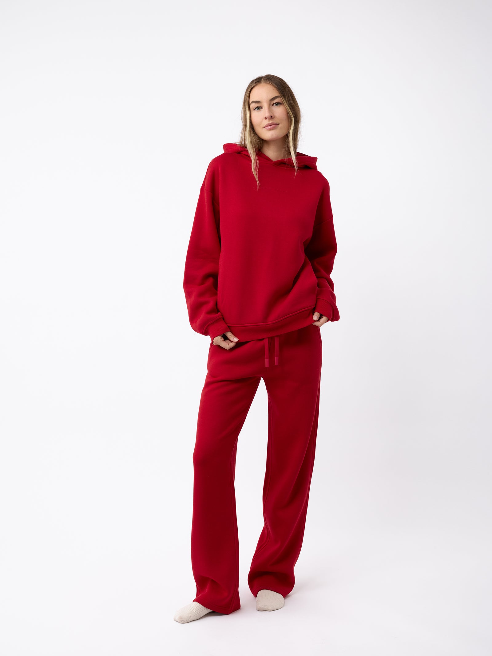 A person stands against a white background wearing a matching red hoodie and Cozy Earth's Women's CityScape Wide Leg Pant. They have long hair and are barefoot, with hands in the hoodie pockets, exuding a casual and relaxed vibe. 