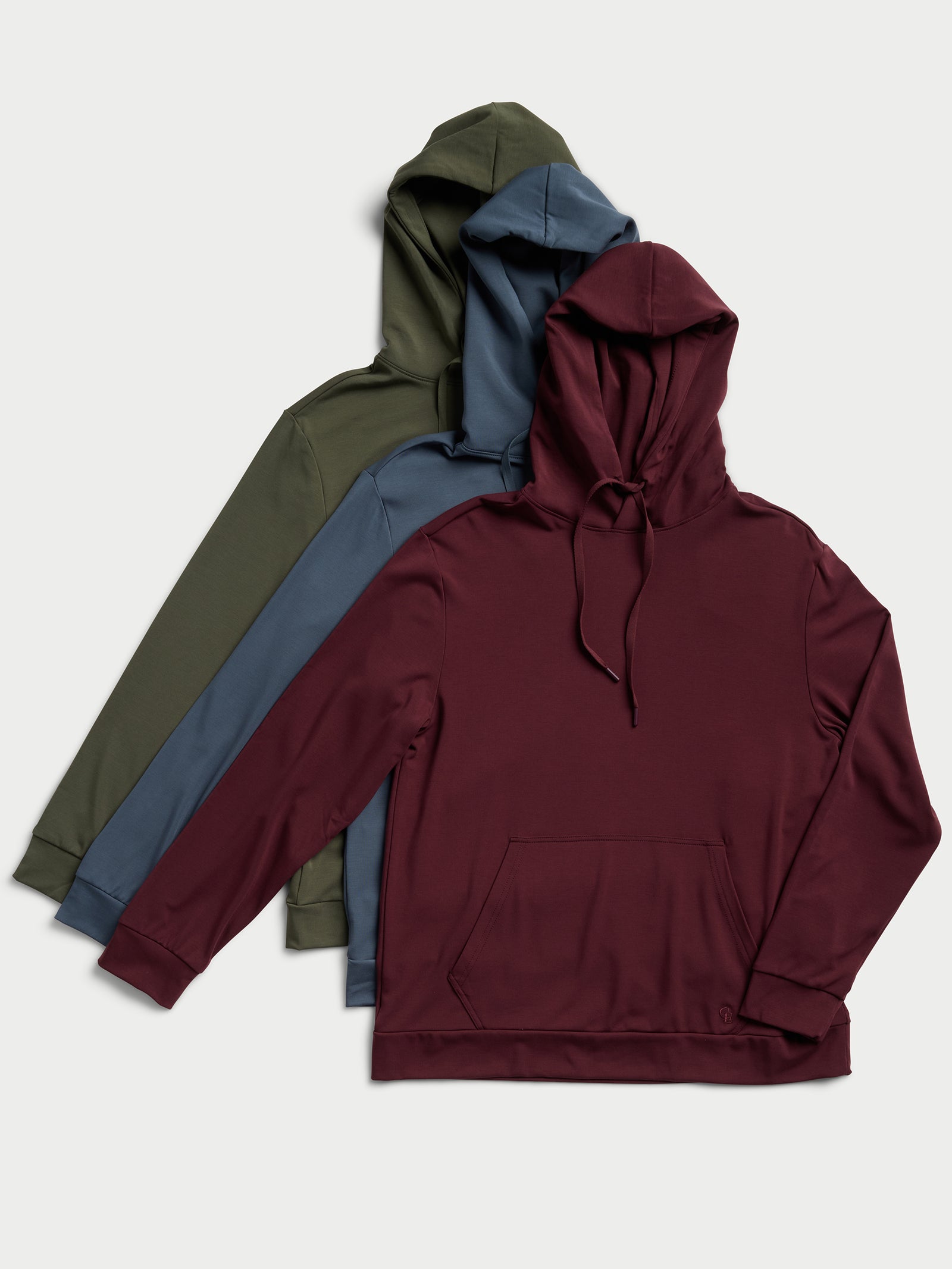 Three Men's Ultra-Soft Bamboo Hoodies from Cozy Earth are displayed in an overlapping arrangement, highlighting burgundy, teal, and olive green shades. Each pullover includes a drawstring hood and a front pocket, set against a light gray background. 