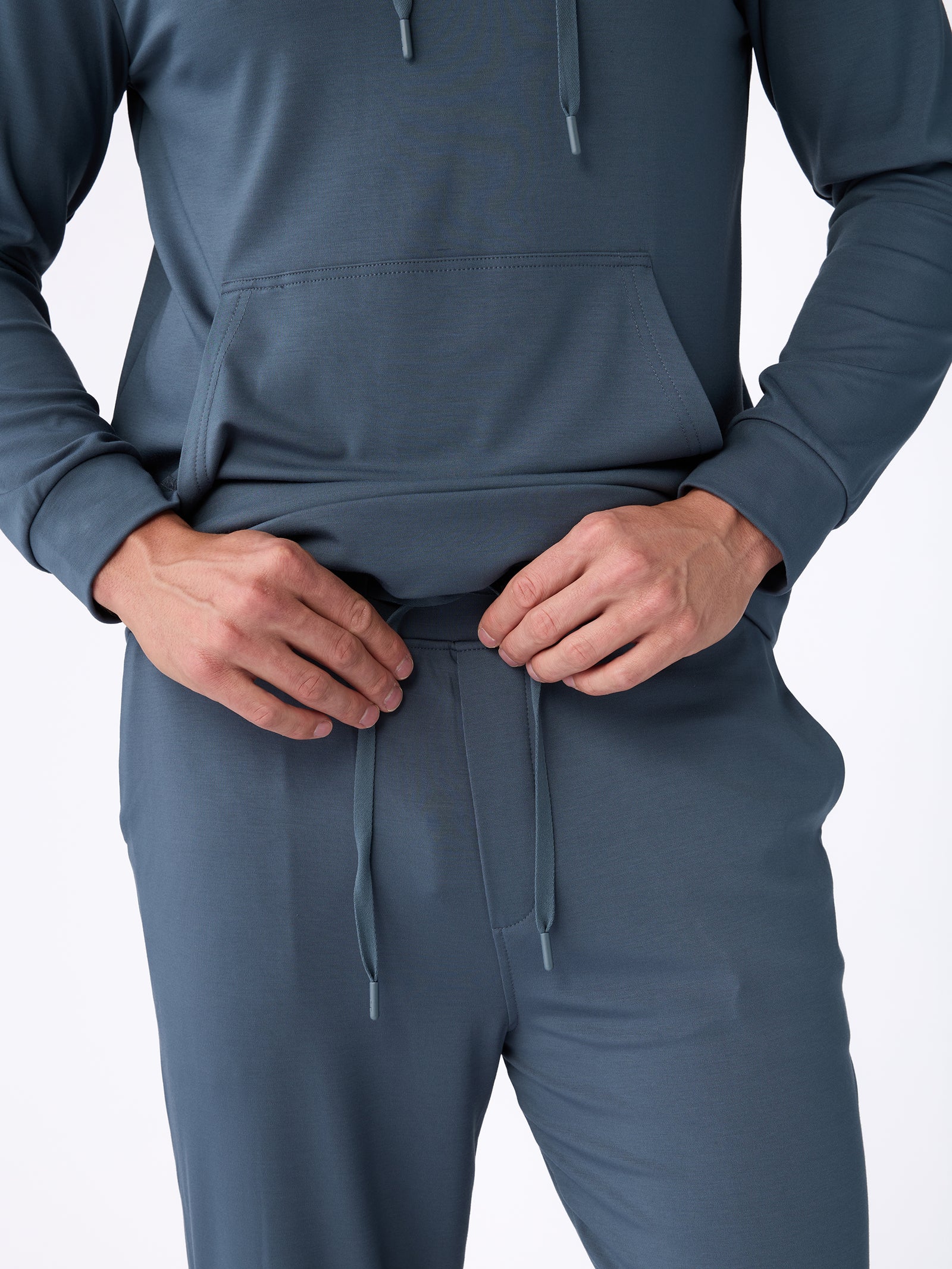 A person wearing a blue tracksuit from Cozy Earth's Men's Ultra-Soft Bamboo Pullover Crew collection adjusts the waistband's drawstring. The outfit includes a hooded sweatshirt with a kangaroo pocket and matching pants, shown against a plain white background. 