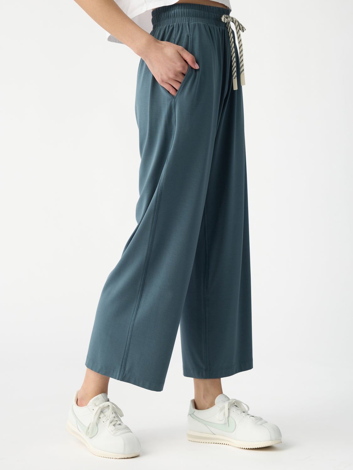 A person wears the Women's Studio Cropped Wide Leg Pant by Cozy Earth in teal, with hands in pockets and paired with white sneakers. The pants feature an elastic waistband and striped drawstring. The photo is cropped above the torso against a white background. 