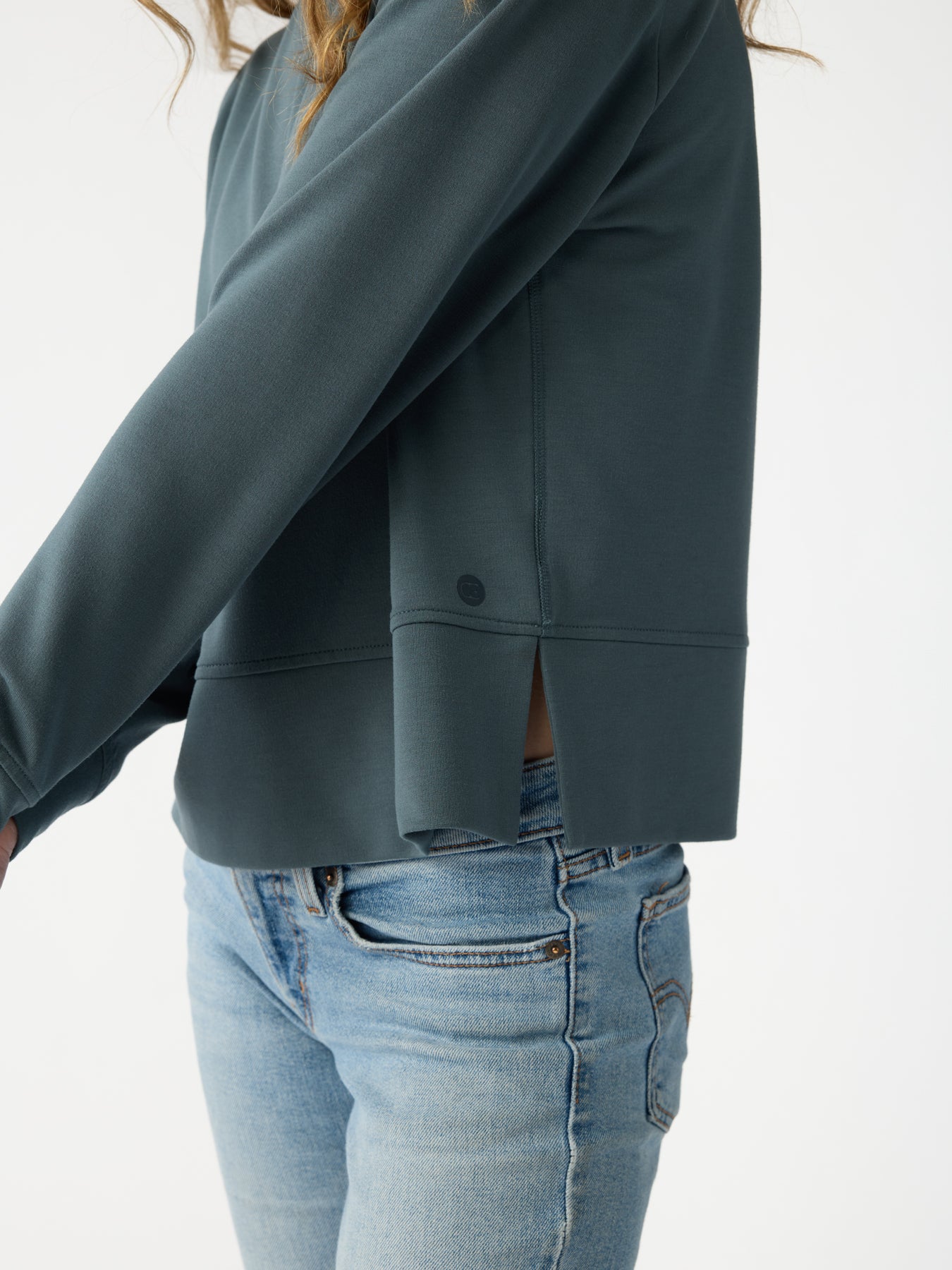 A person is wearing the Cozy Earth Women's StudioLite Crewneck, a dark green long-sleeve top with side split detail, paired with light blue jeans against a plain background. The focus is on the outfit, highlighting the texture and fit of the clothing. 