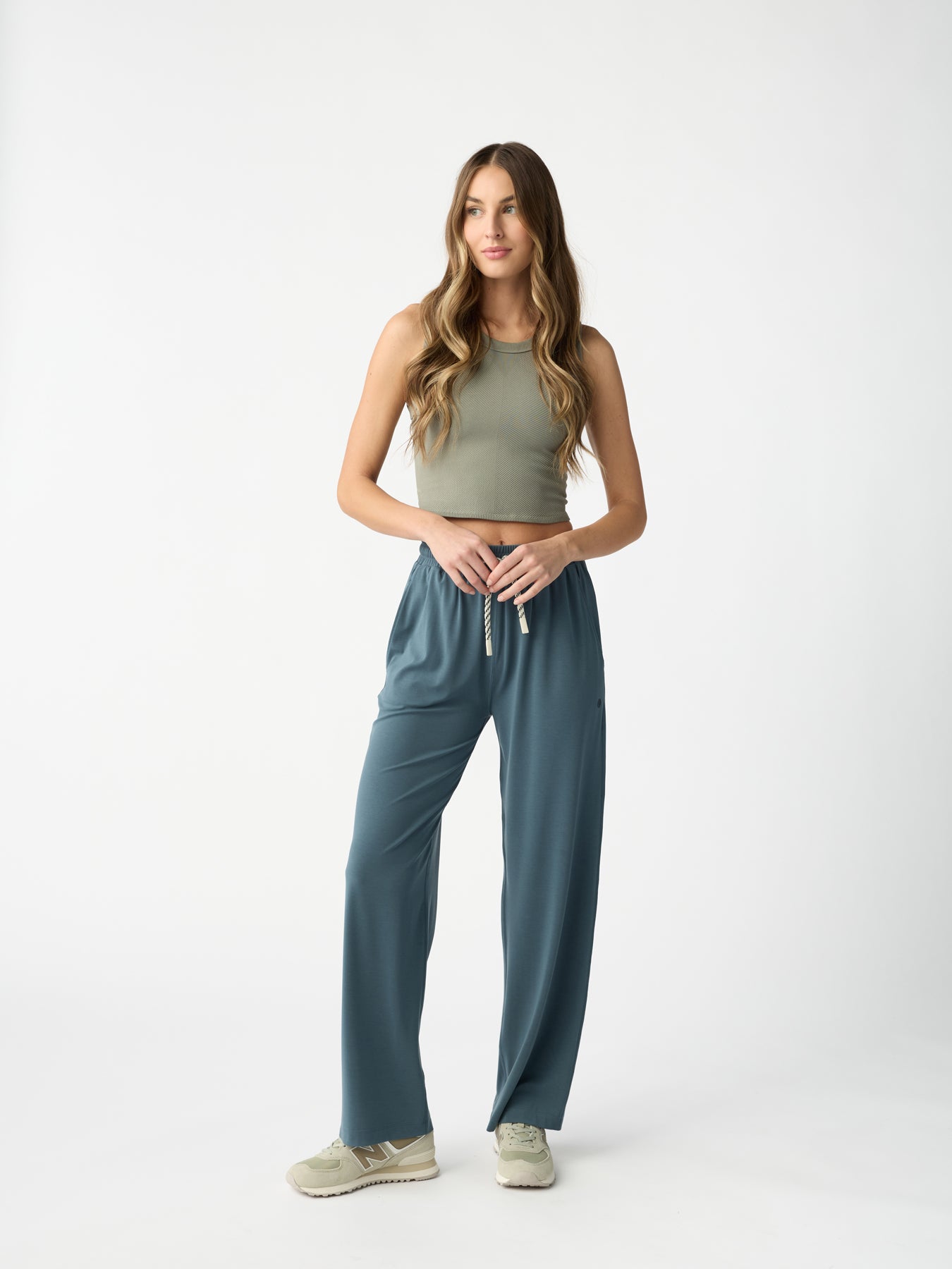 A woman with long hair poses against a plain white background, wearing an olive green sleeveless top, Cozy Earth's Women's Studio Wide Leg Pant in teal, and beige sneakers. Her hands are in her pockets, and she appears relaxed. 