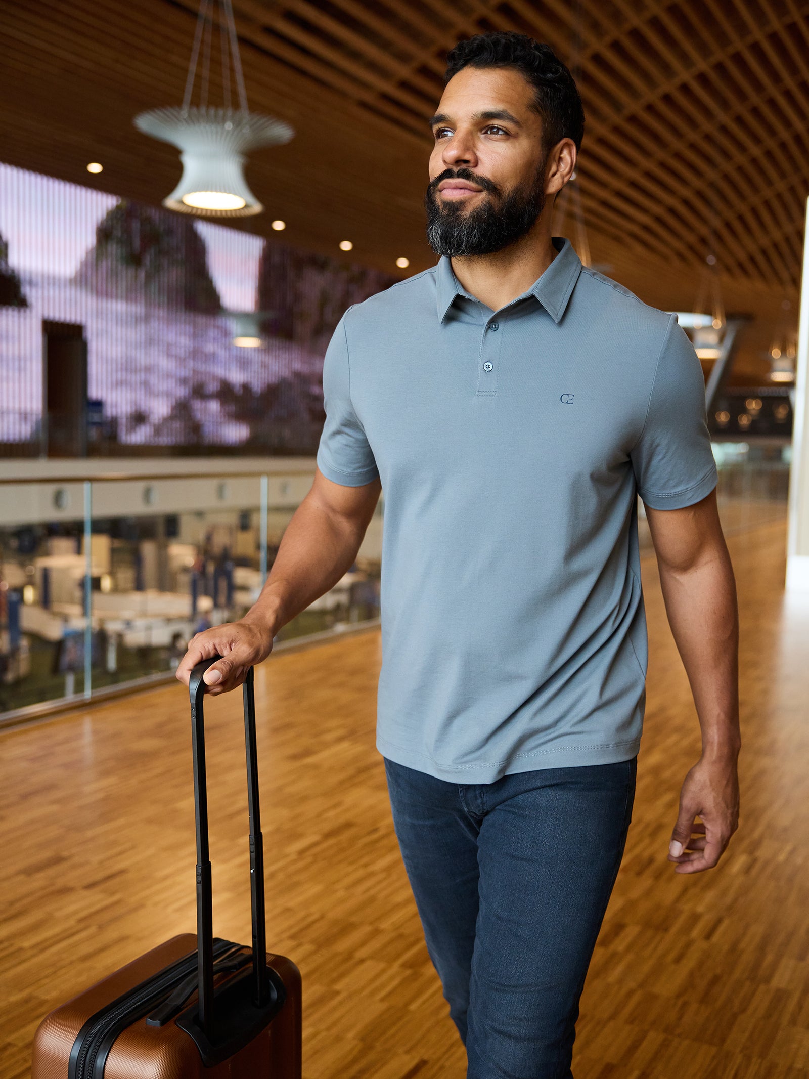 A bearded man in a Men's Everyday Polo by Cozy Earth is walking through an airport terminal, pulling a brown carry-on suitcase. The terminal features wooden floors, a large digital display, and modern overhead lighting. 