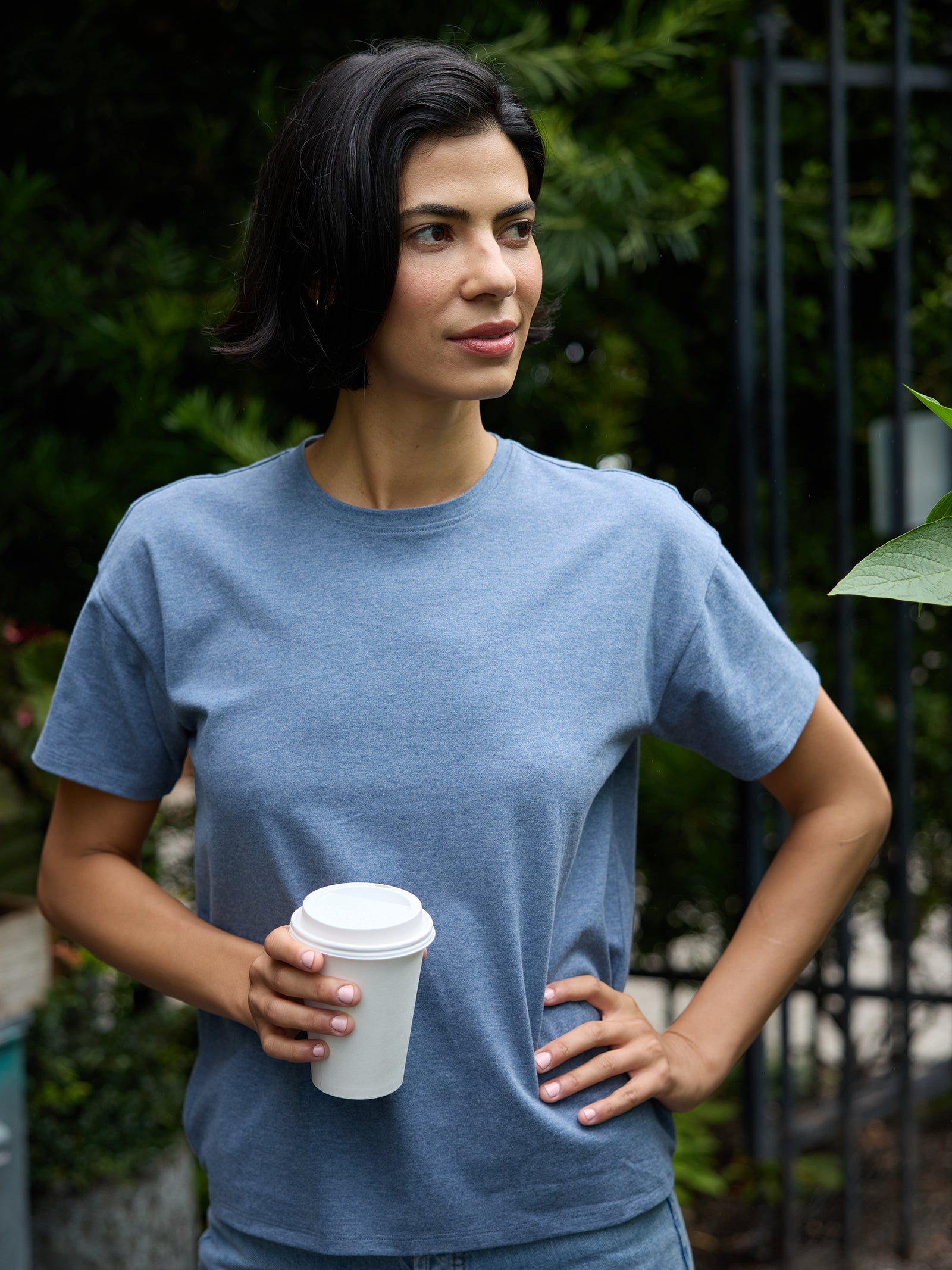 A person with short dark hair stands outdoors, holding a white paper coffee cup in one hand and resting the other hand on their hip. They are wearing a gray Cozy Earth Women's All Day Tee and looking off to the side, with greenery and a metal fence in the background. 